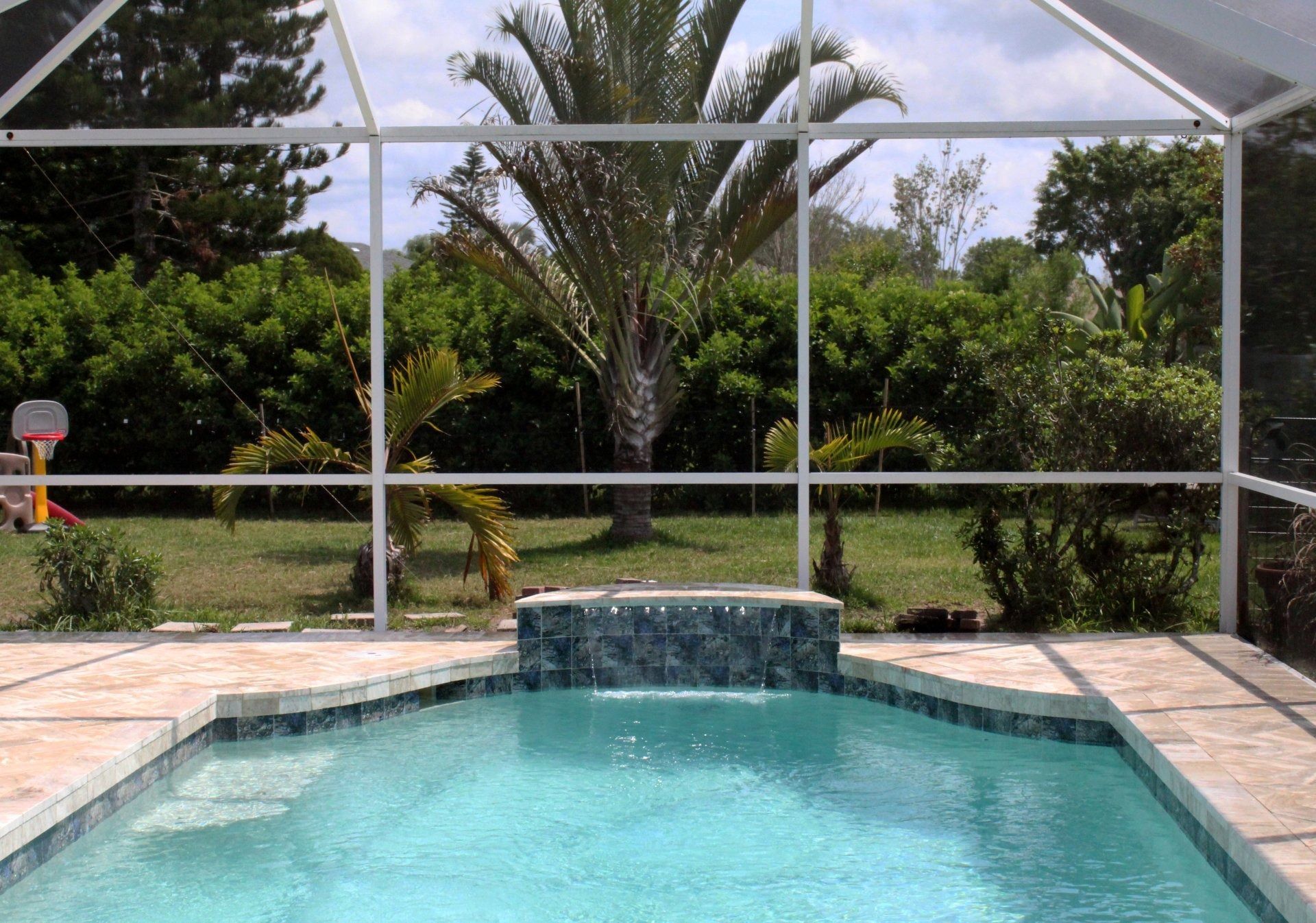 A clear blue swimming pool with a tiled fountain, enclosed by a screen cage overlooking a backyard with palm trees.