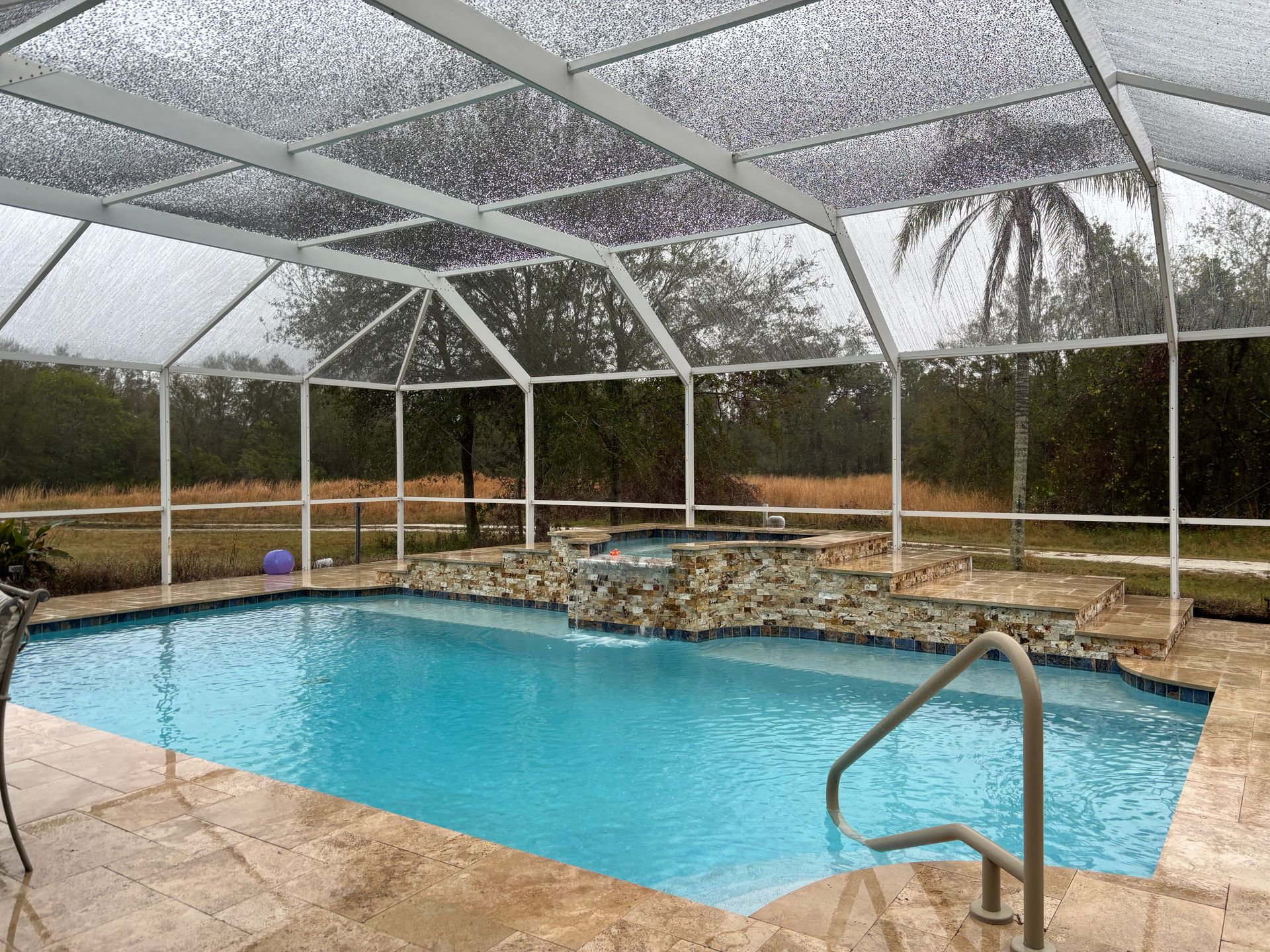 A bright blue rectangular swimming pool with a stone water feature under a white screened enclosure on a rainy day.
