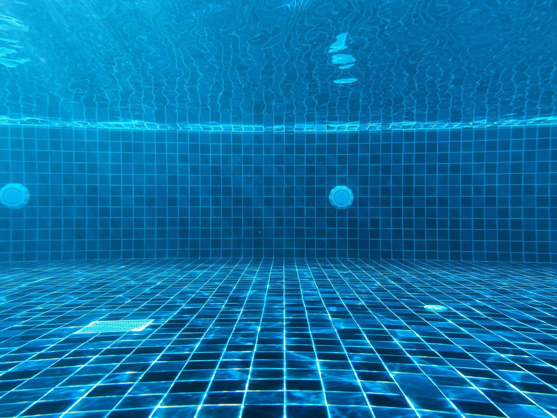 Underwater view of a swimming pool with blue tiled floors and walls, with sunlight filtering through the water surface.