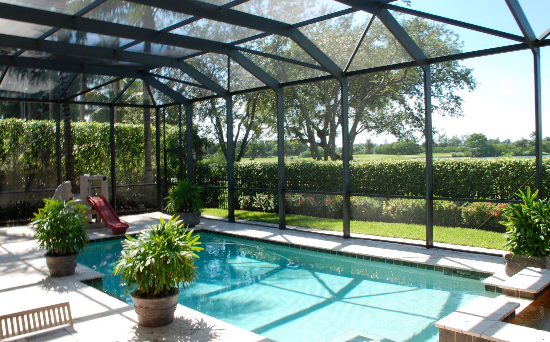 A swimming pool inside a screened-in lanai, overlooking a lush green lawn and trees on a sunny day.