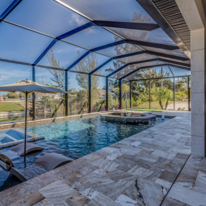 A screen-enclosed patio with a swimming pool, tanning ledges, a spa, and beige stone tiles overlooking a grassy lawn.