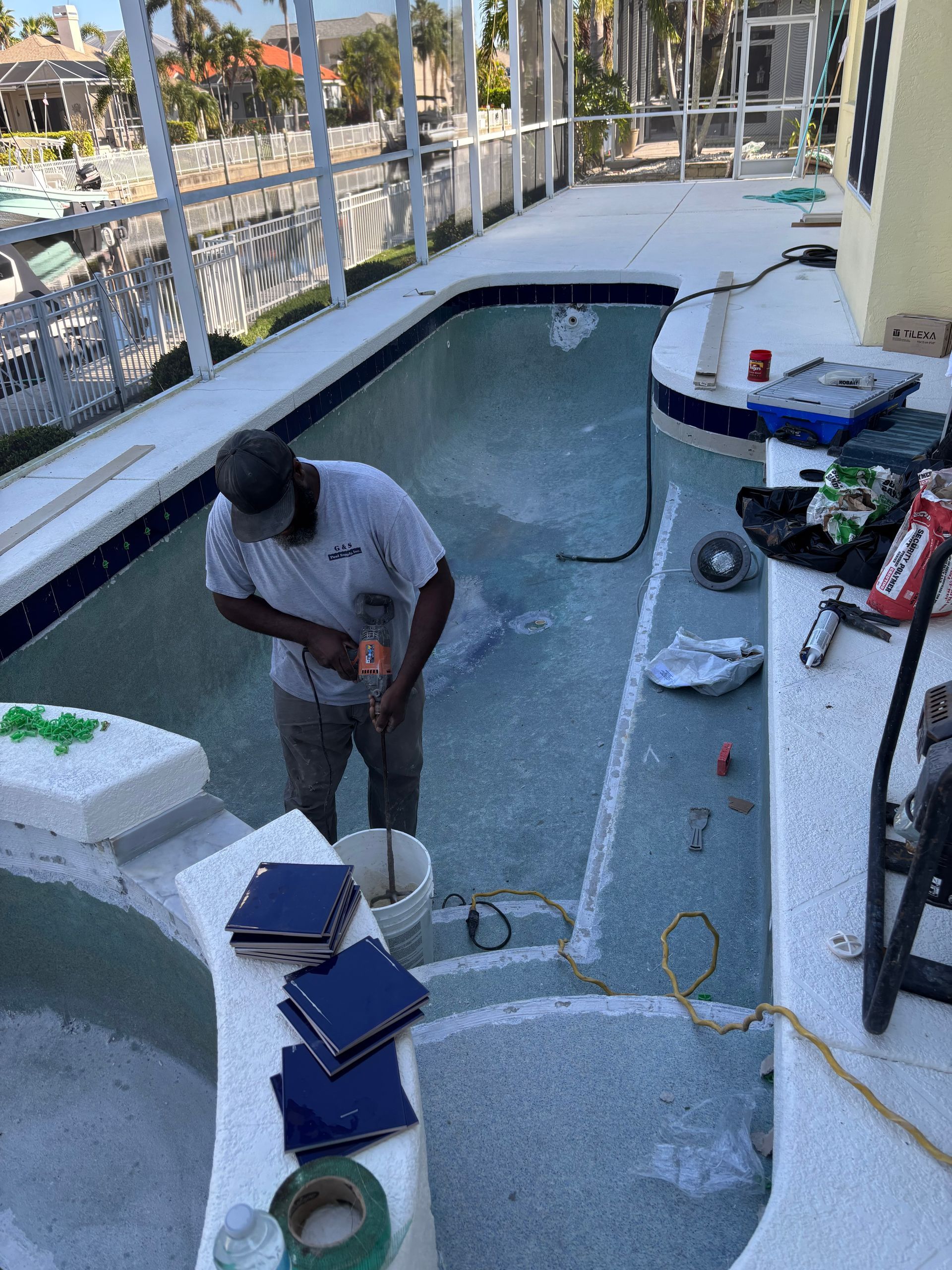 A worker uses a power tool to repair the edge of a light-colored swimming pool inside a screened enclosure.