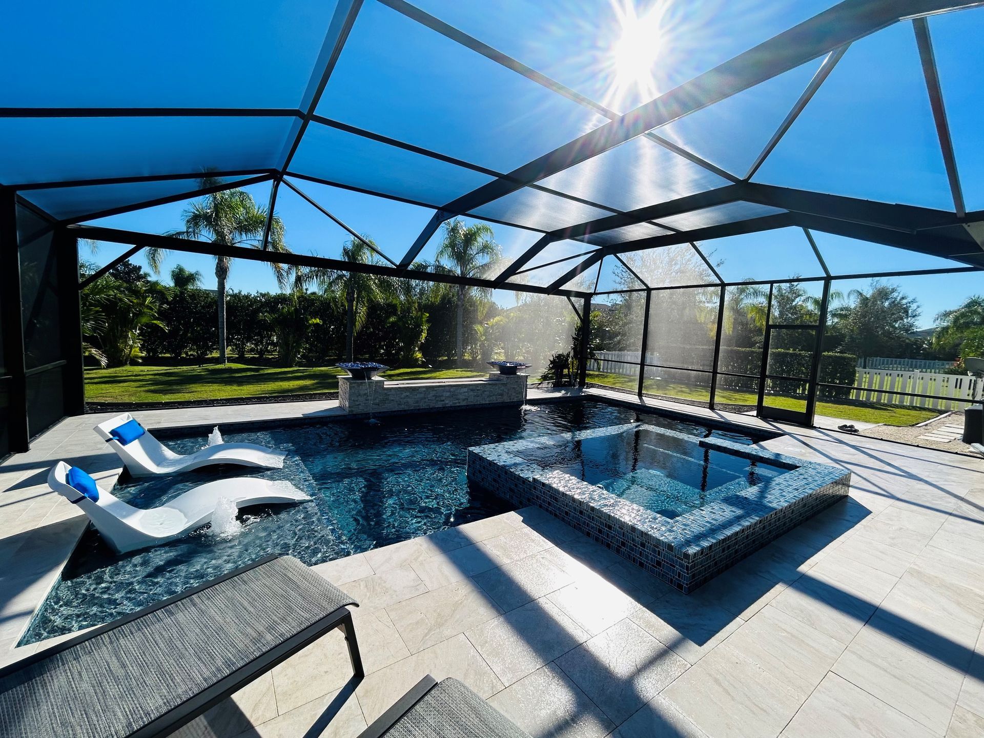 A bright outdoor swimming pool with a connected hot tub, featuring lounge chairs under a black screened-in enclosure.
