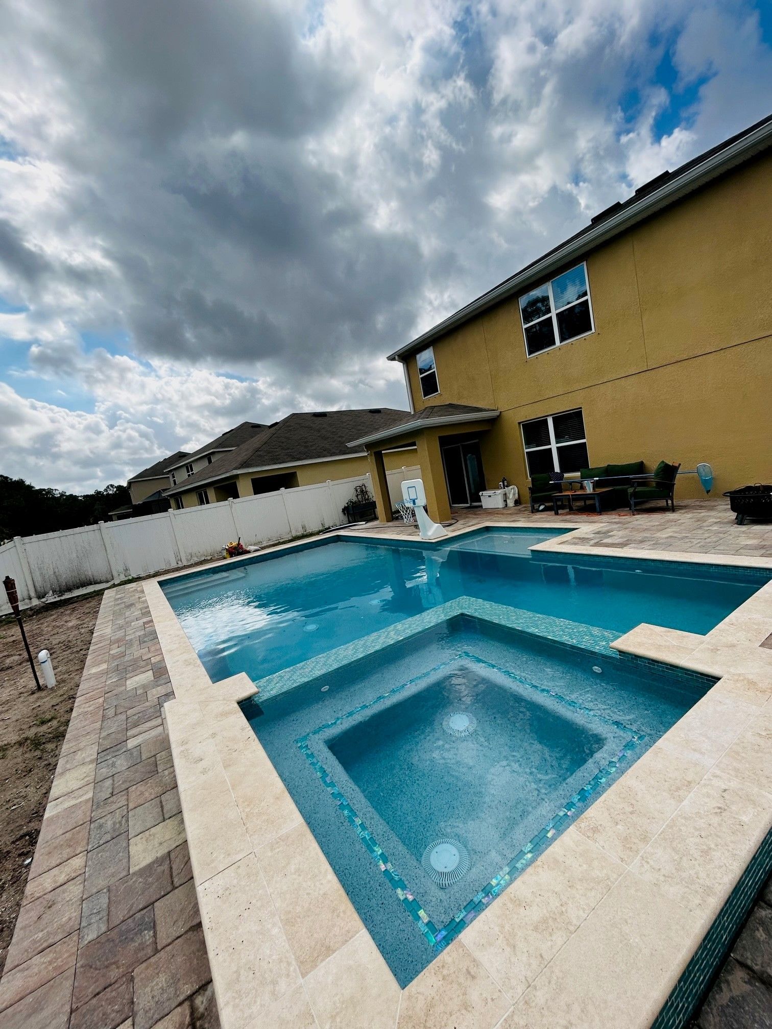An in-ground swimming pool and hot tub with light tan pavers next to a two-story yellow house under a cloudy sky.