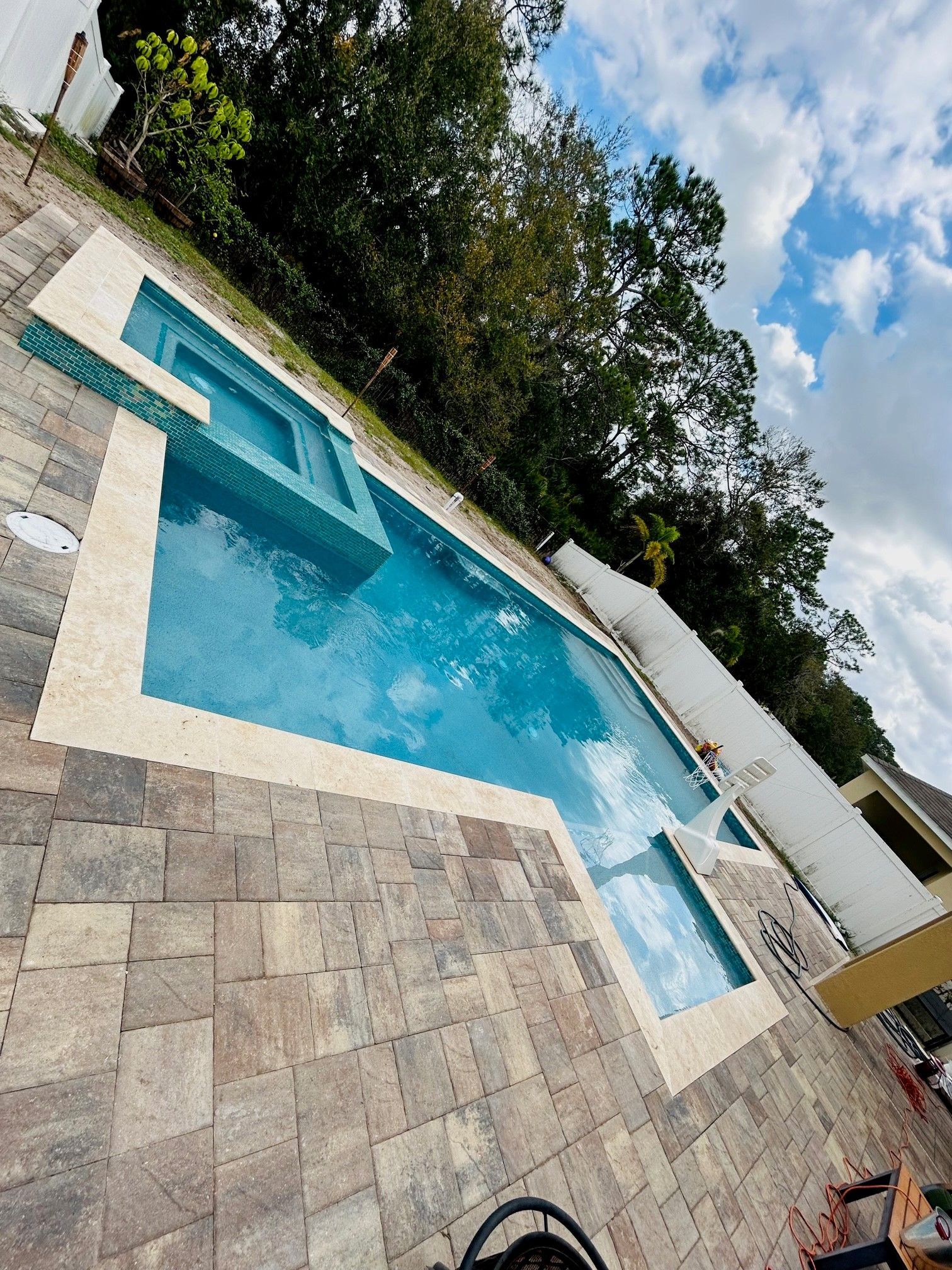 A rectangular backyard pool with an attached spa, surrounded by stone pavers, trees, and a white fence under a cloudy sky.