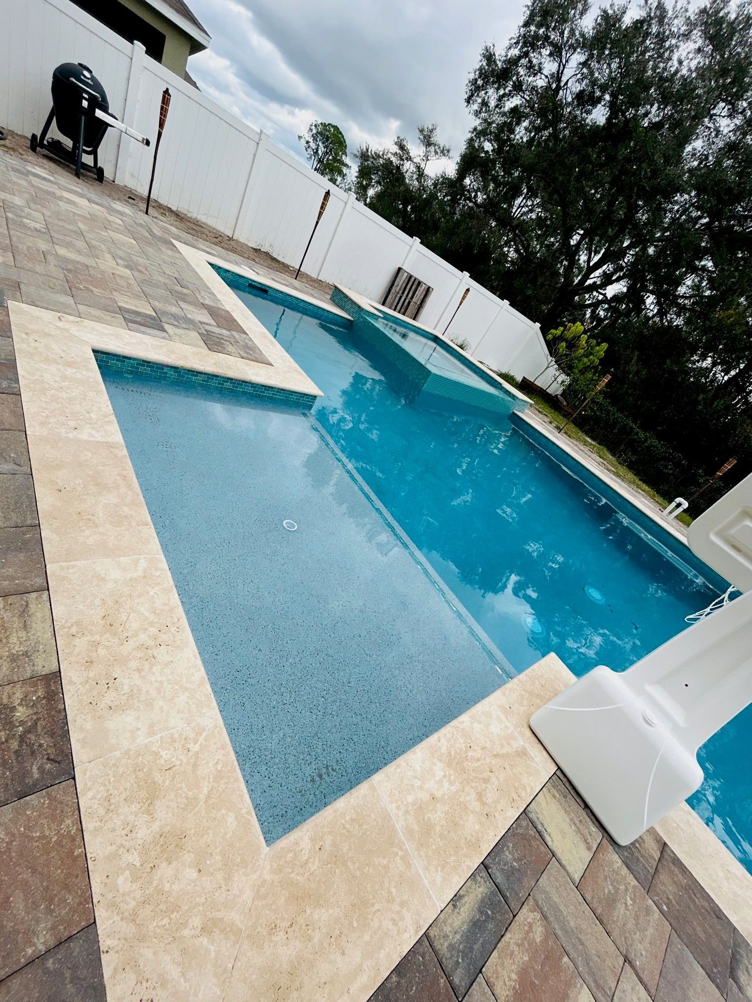 A backyard swimming pool with a stone deck, a raised spa, and a white privacy fence against a backdrop of trees.
