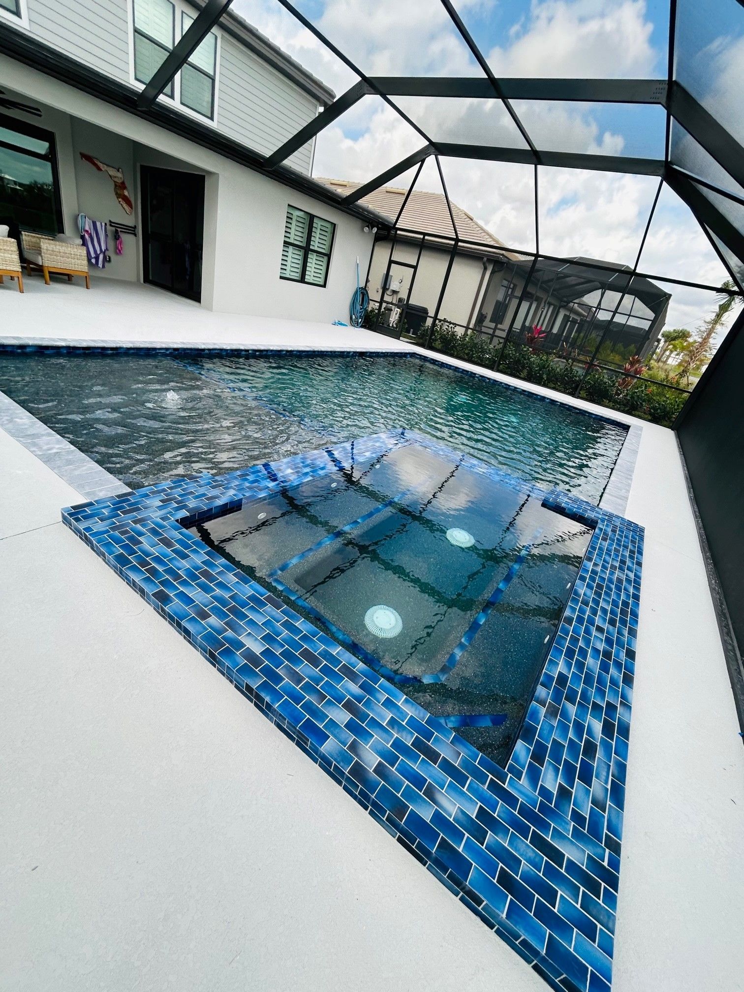 A screened-in backyard pool and hot tub with deep blue glass tiles, light-colored deck, and adjacent house wall.