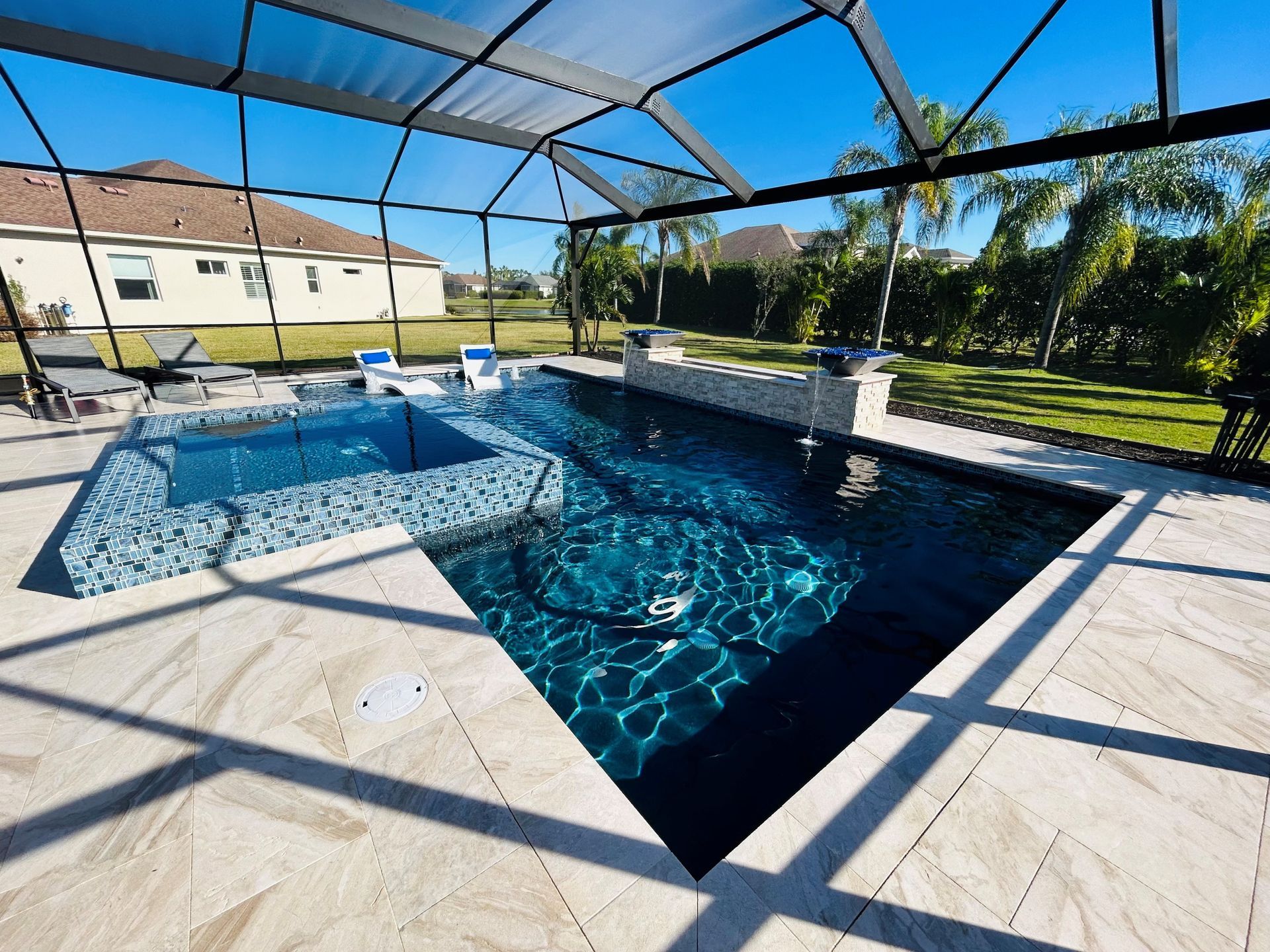 A rectangular pool and raised spa with blue mosaic tiles, set on a light stone patio under a screened enclosure.