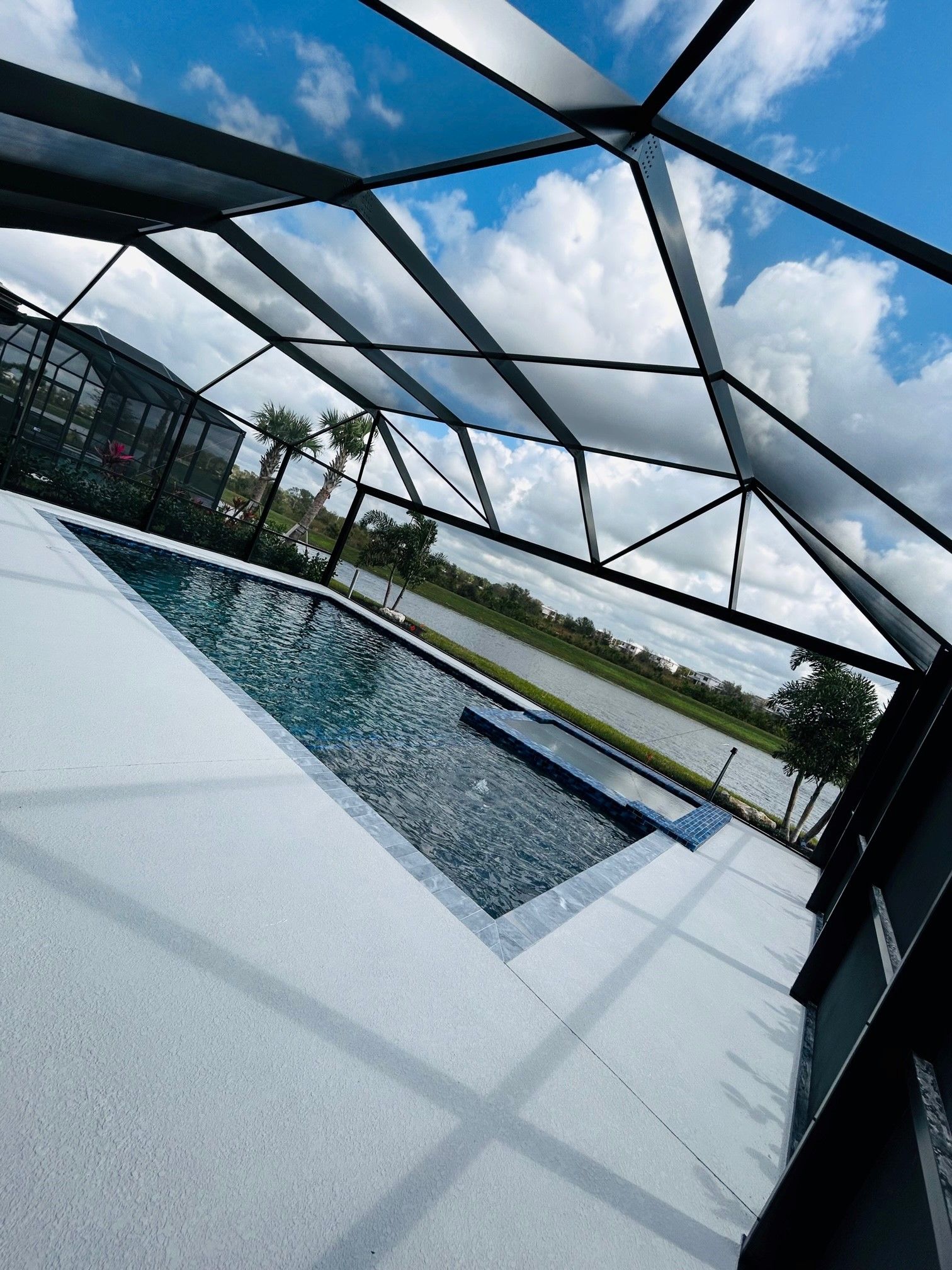 A swimming pool under a screened enclosure overlooking a grassy yard and a pond under a cloudy blue sky.