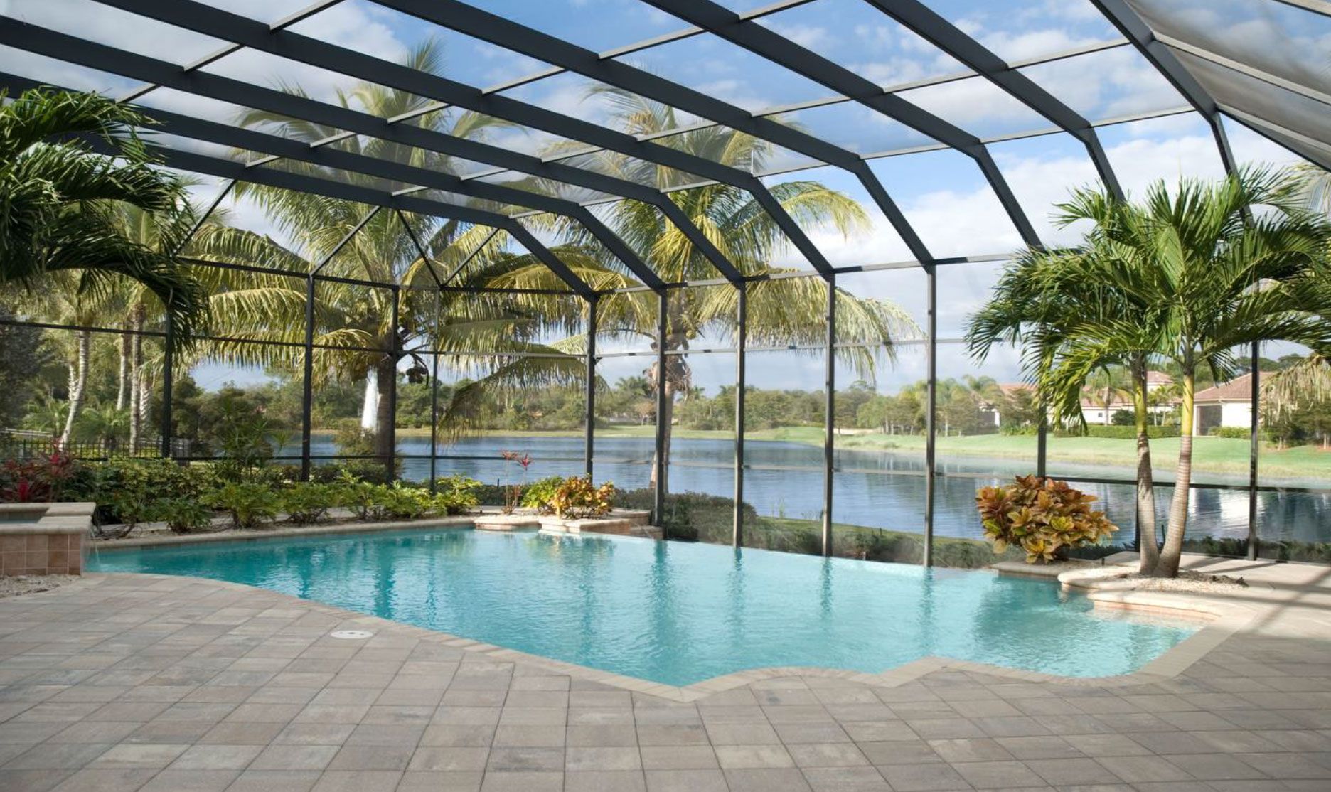 A screened-in swimming pool patio overlooking a calm lake surrounded by palm trees and lush greenery.
