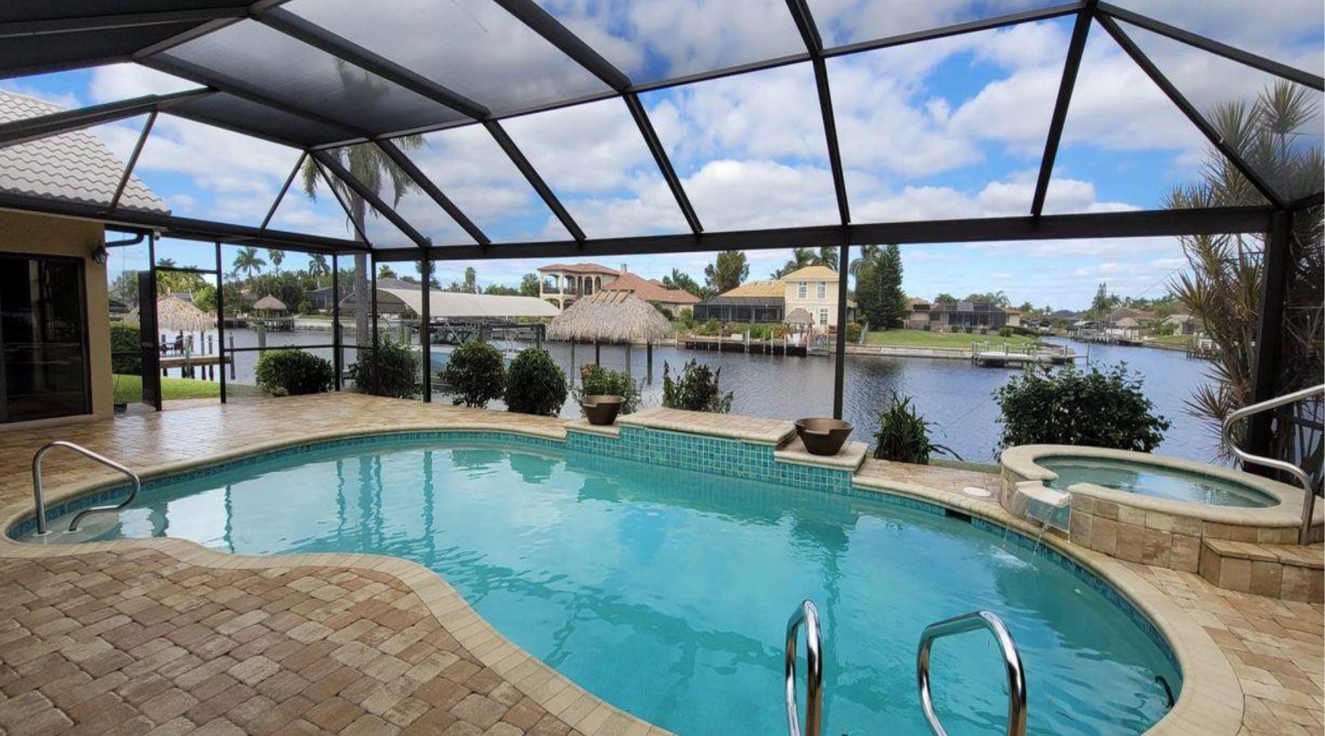 Screen-enclosed swimming pool and spa overlooking a canal with houses in the background under a partly cloudy sky.