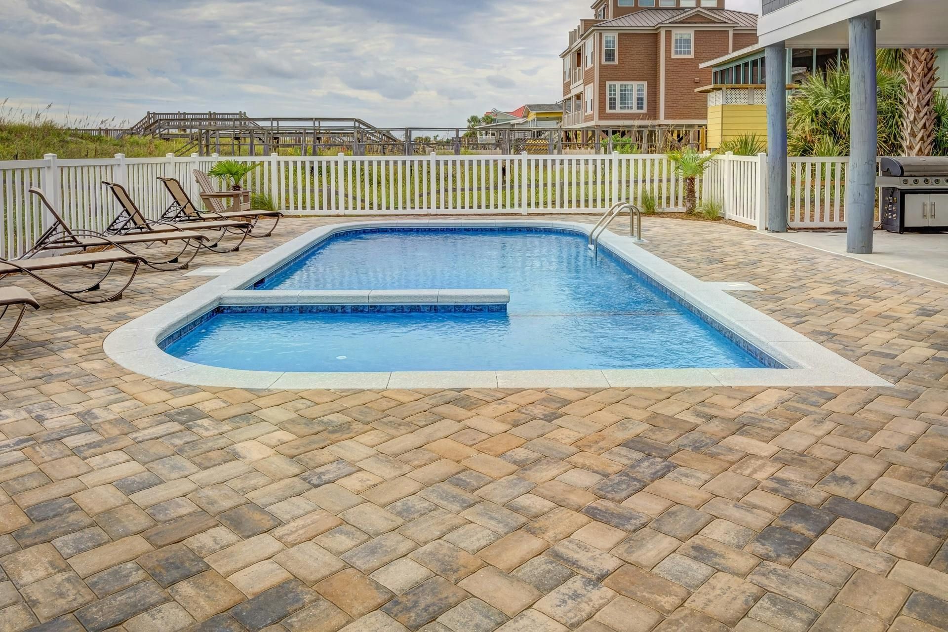 A blue, L-shaped swimming pool sits in a stone-paved patio area with lounge chairs, a white fence, and a house nearby.