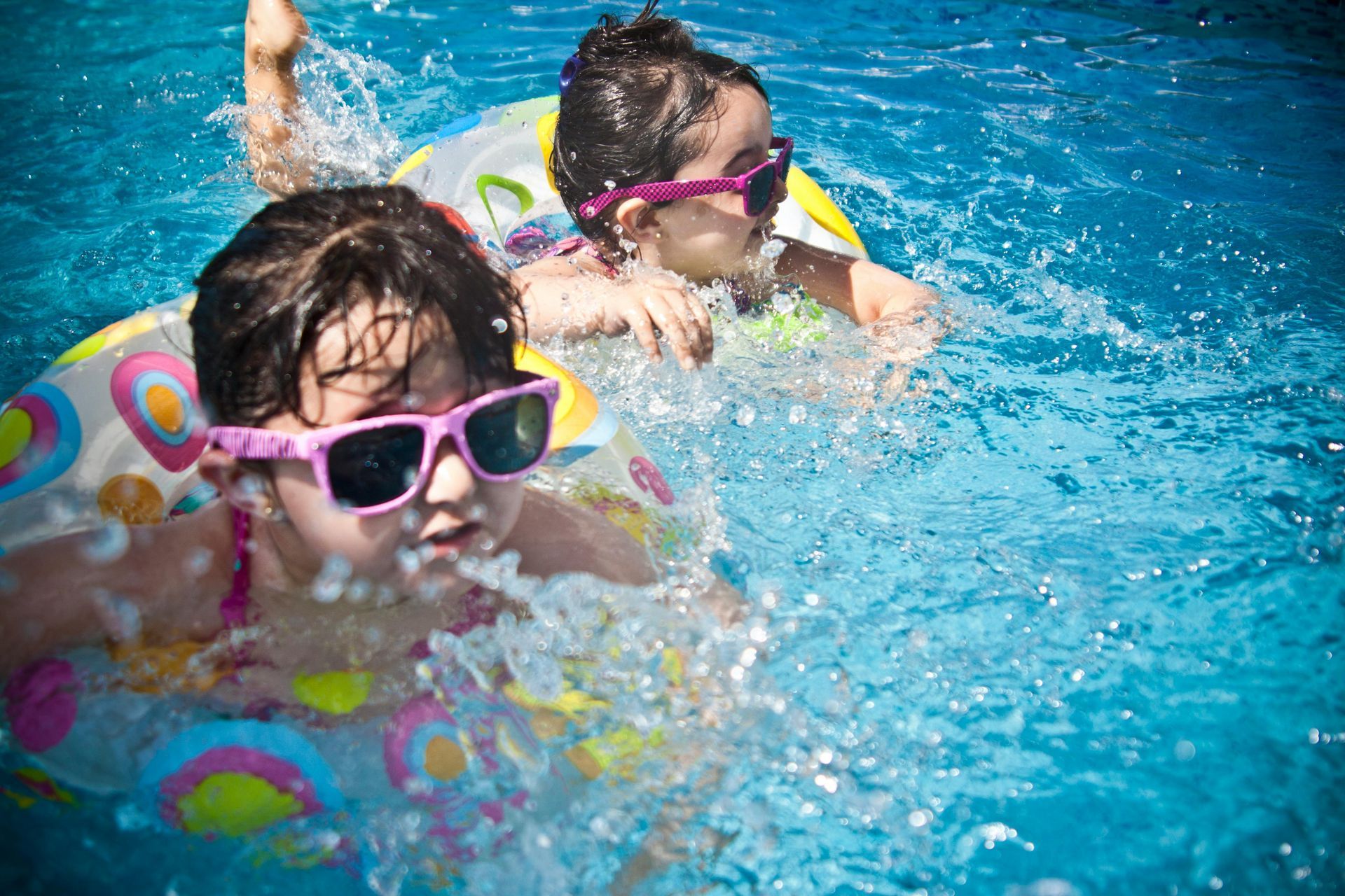 Two children wearing sunglasses play in a swimming pool using colorful inflatable rings.