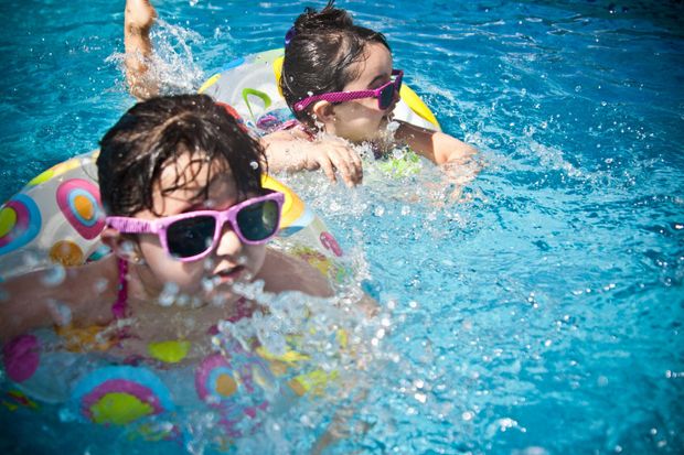 Two people in a pool, wearing pink sunglasses and using colorful patterned swim rings, surrounded by water splashes.