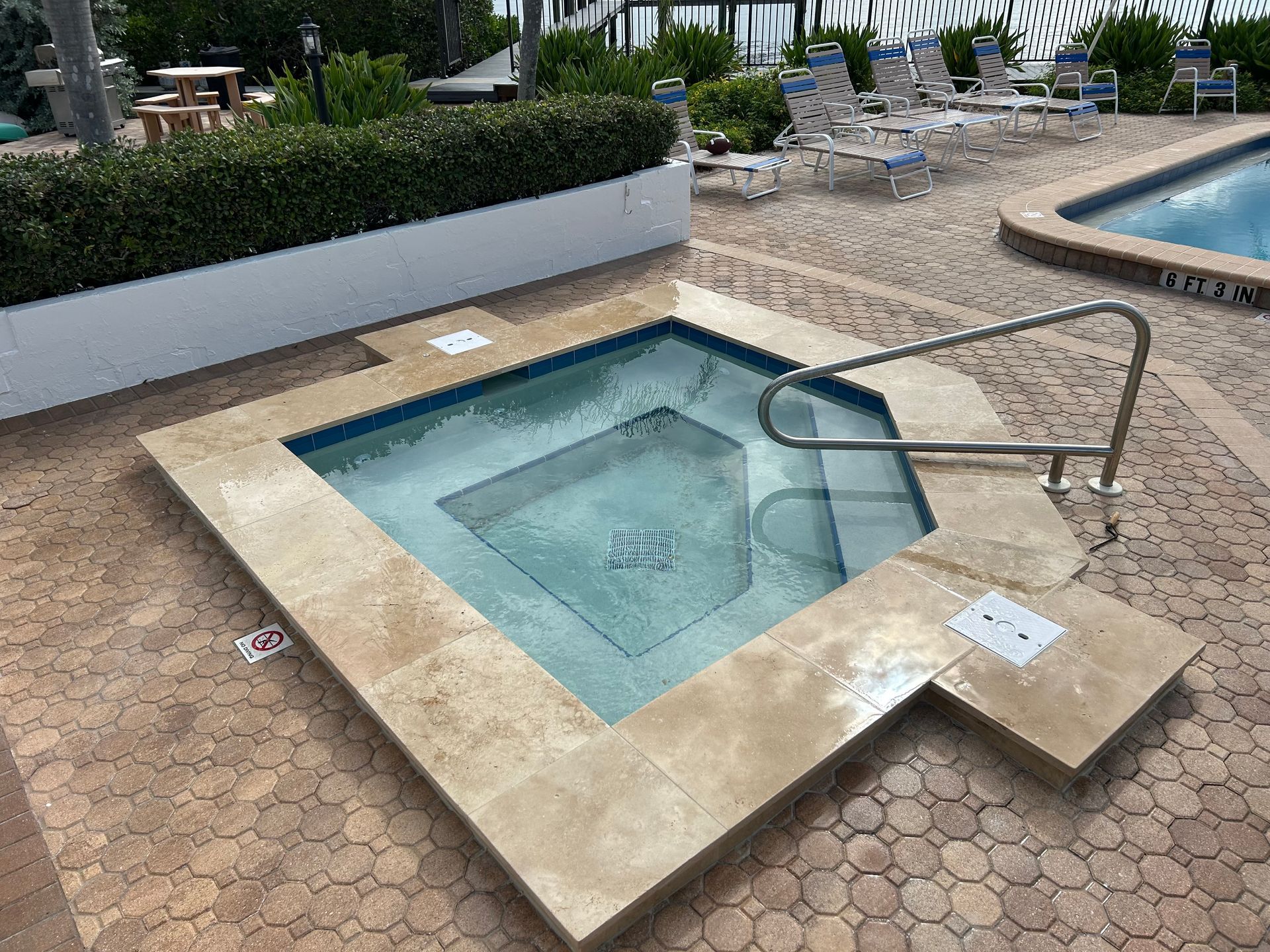 A raised square hot tub with beige stone coping, a metal handrail, and blue tiled interior on a stone pool deck.