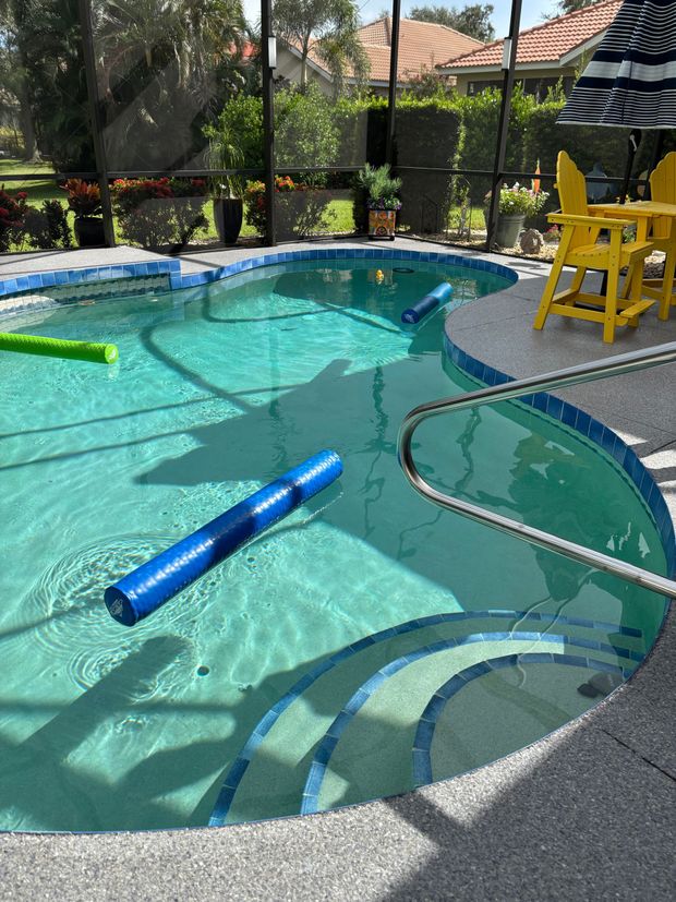 A sunlit swimming pool with blue steps, floating pool noodles, and yellow chairs on a patio enclosed by a screen.