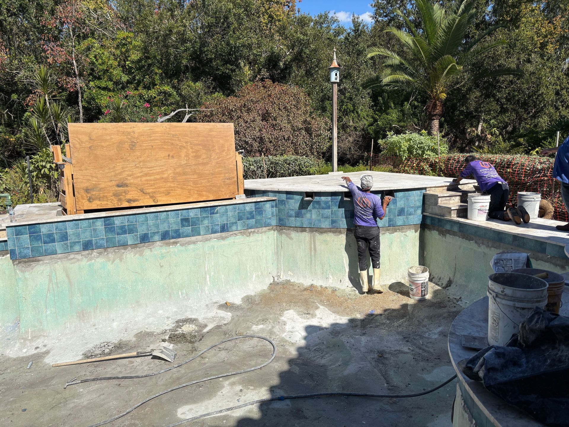 Construction workers renovate a drained swimming pool with blue tiles, standing on a sand floor under a sunny sky.
