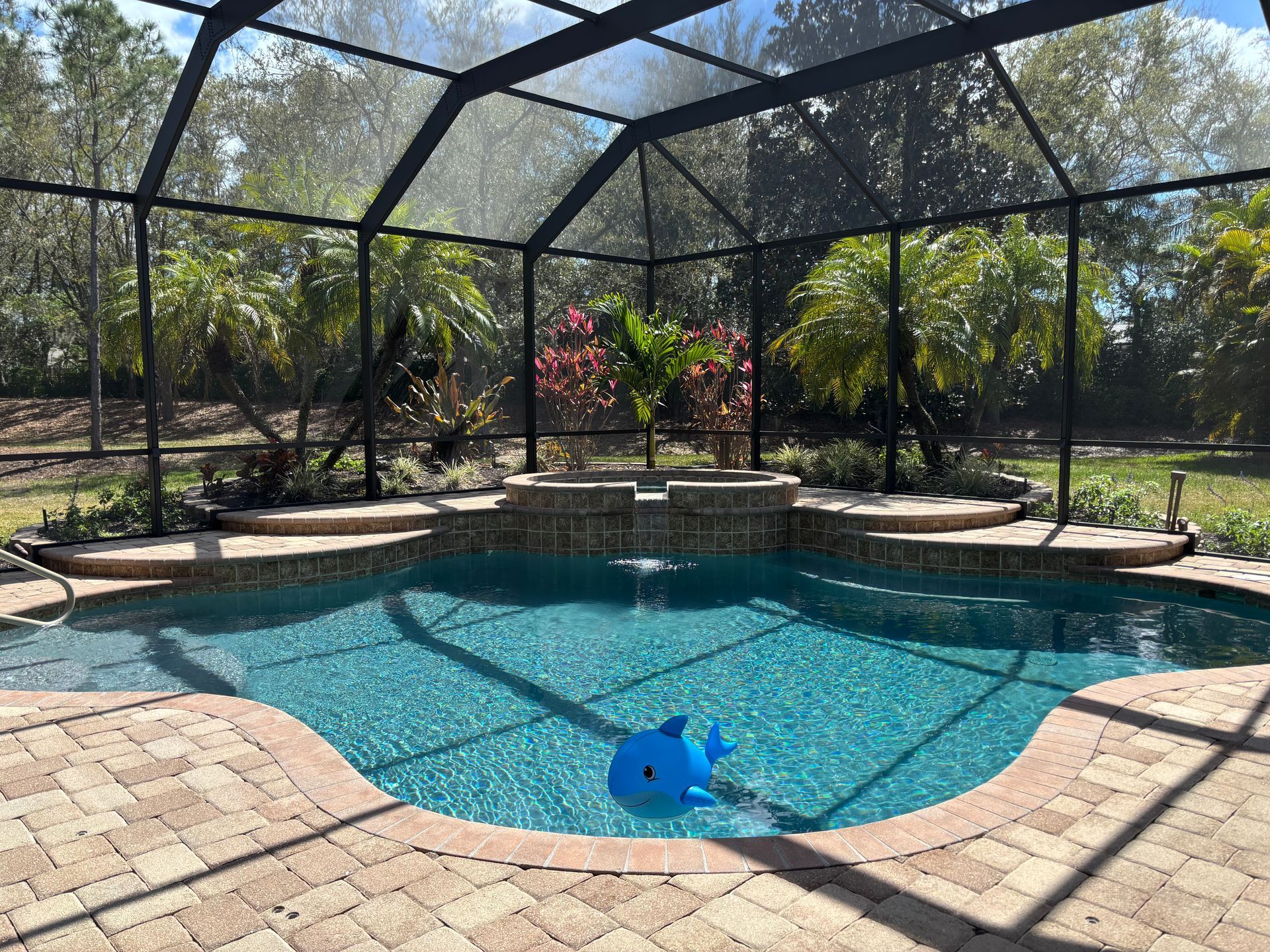 A backyard swimming pool with a raised hot tub and waterfall, enclosed by a screen cage with lush greenery in the back.
