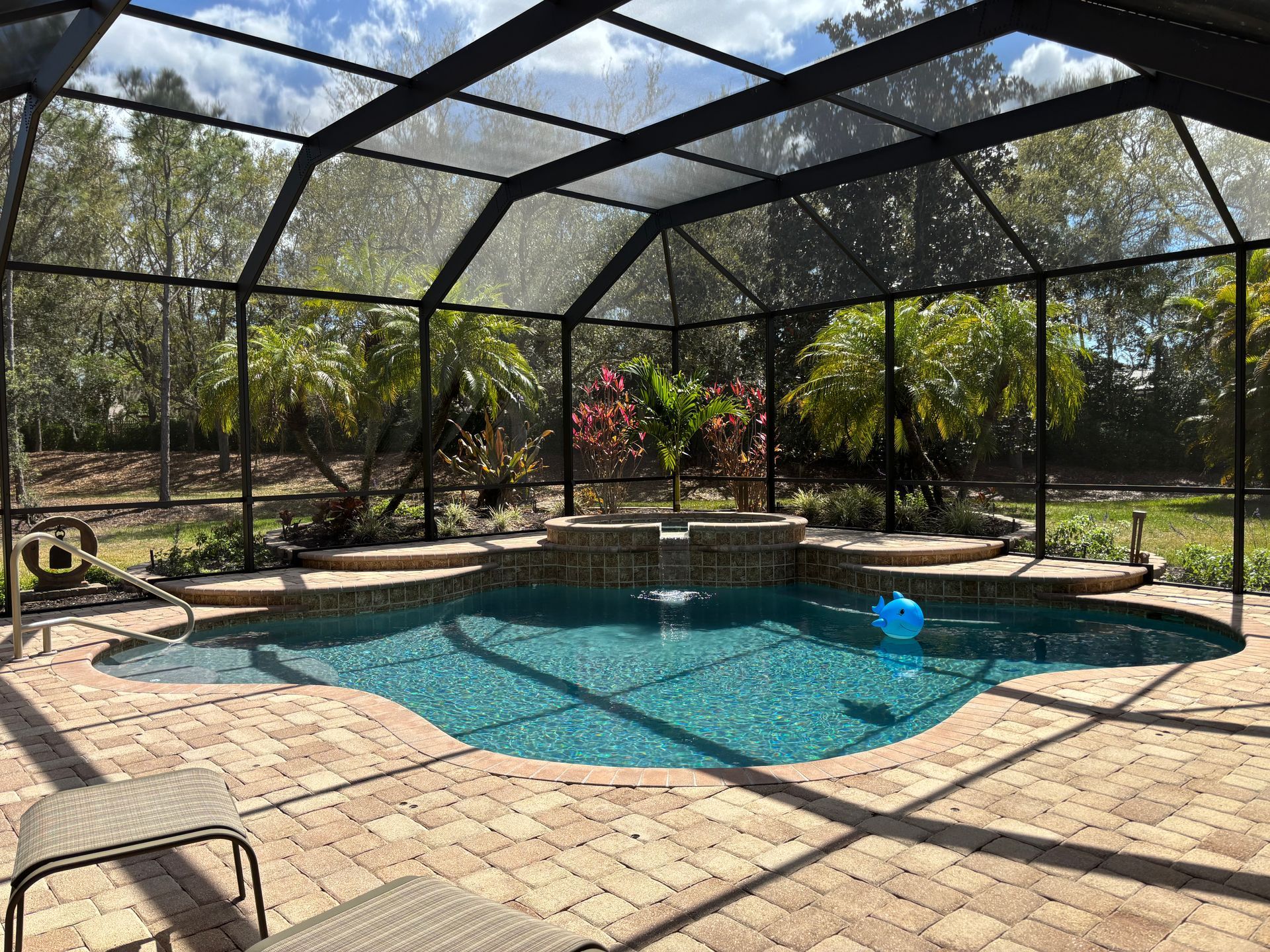 A backyard swimming pool with a stone patio enclosed by a screened-in lanai, surrounded by trees and landscaping.