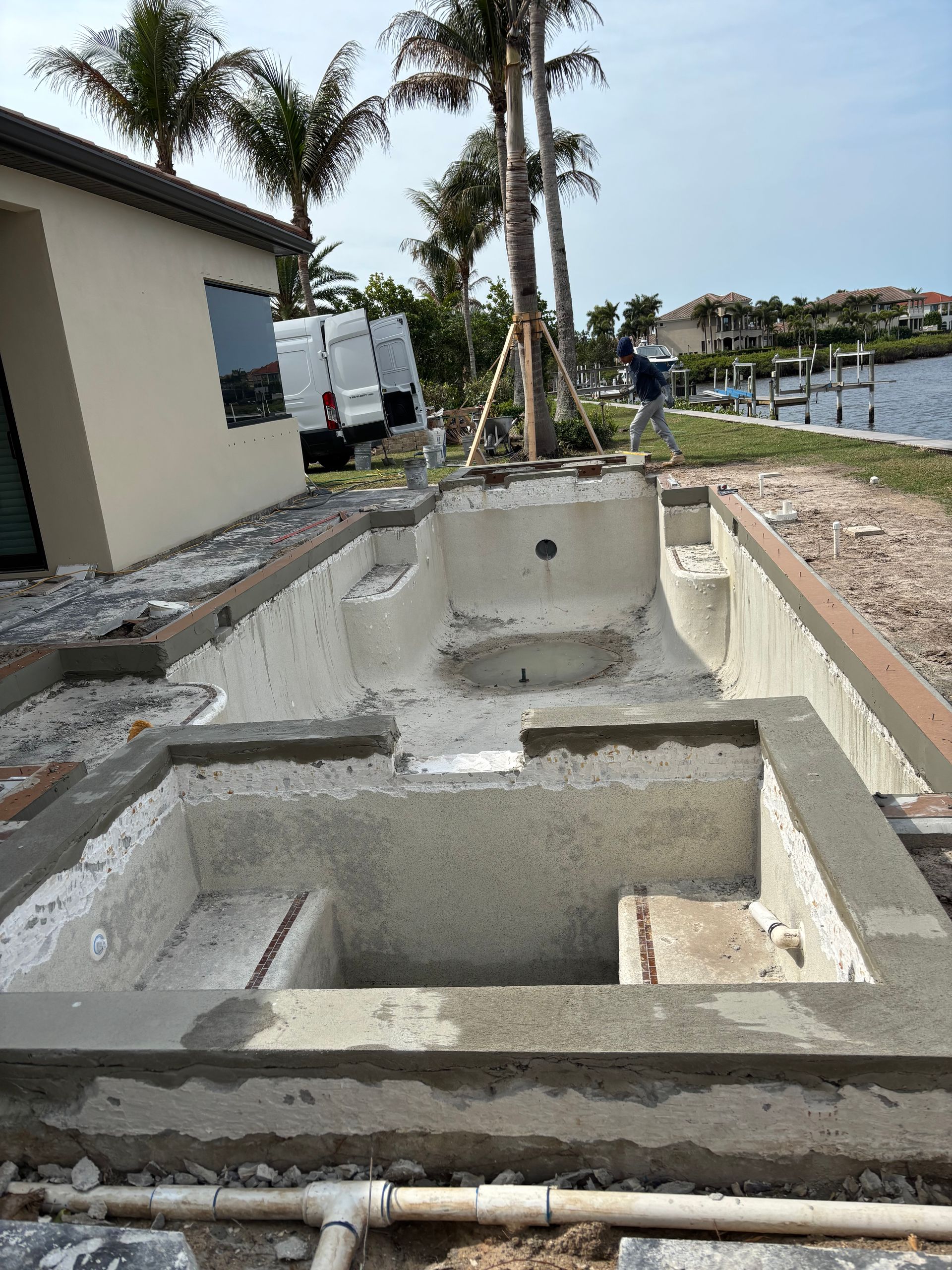 A swimming pool and spa under construction outside a house, with concrete framing, visible pipes, and waterfront views.