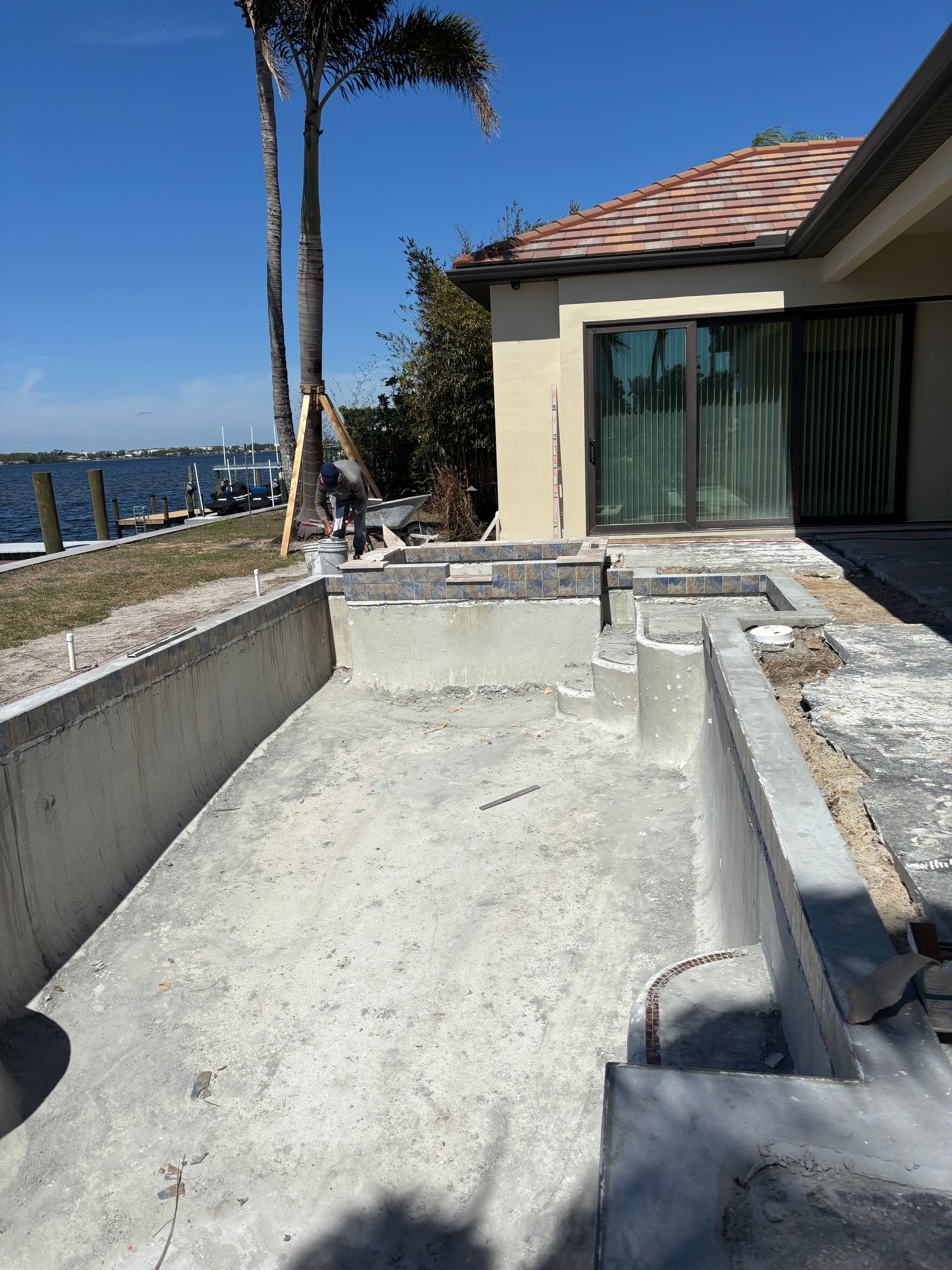 An unfinished concrete swimming pool structure under construction next to a house overlooking a body of water.