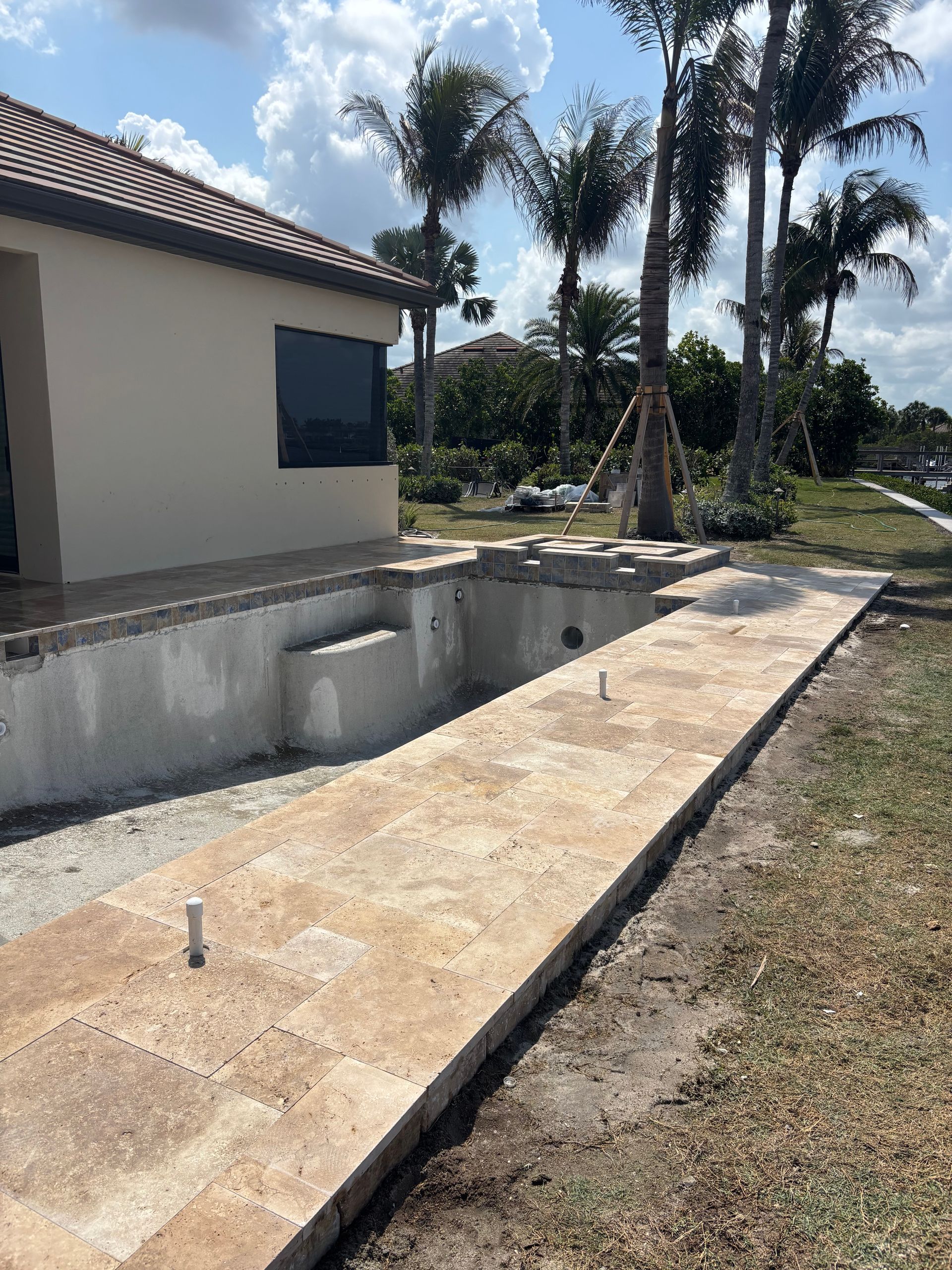 A partially constructed pool with tan stone coping and decking outside a residential home under a blue, cloudy sky.