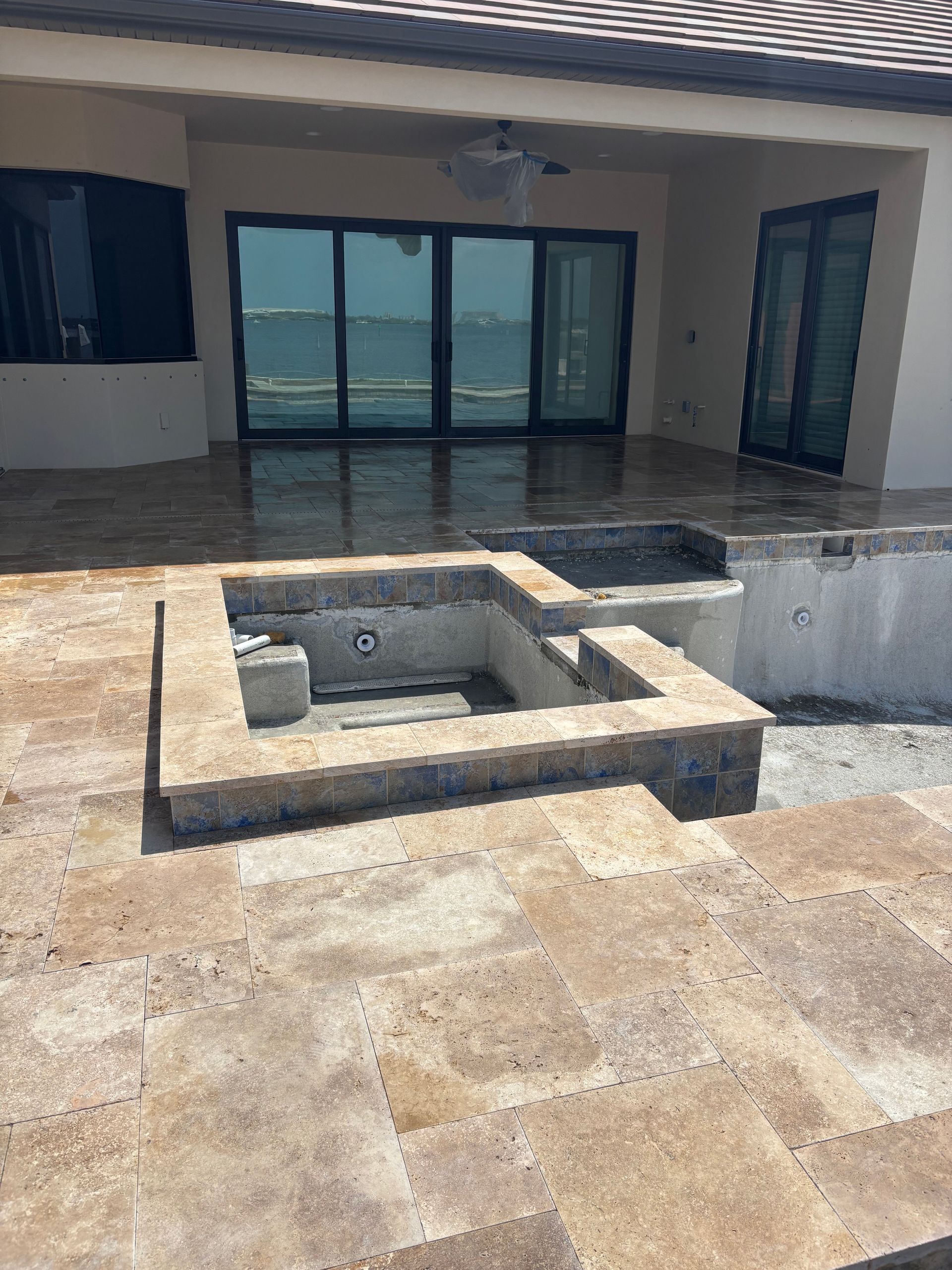 A newly constructed square concrete hot tub spa sits on a beige stone patio outside a home with large sliding glass doors.
