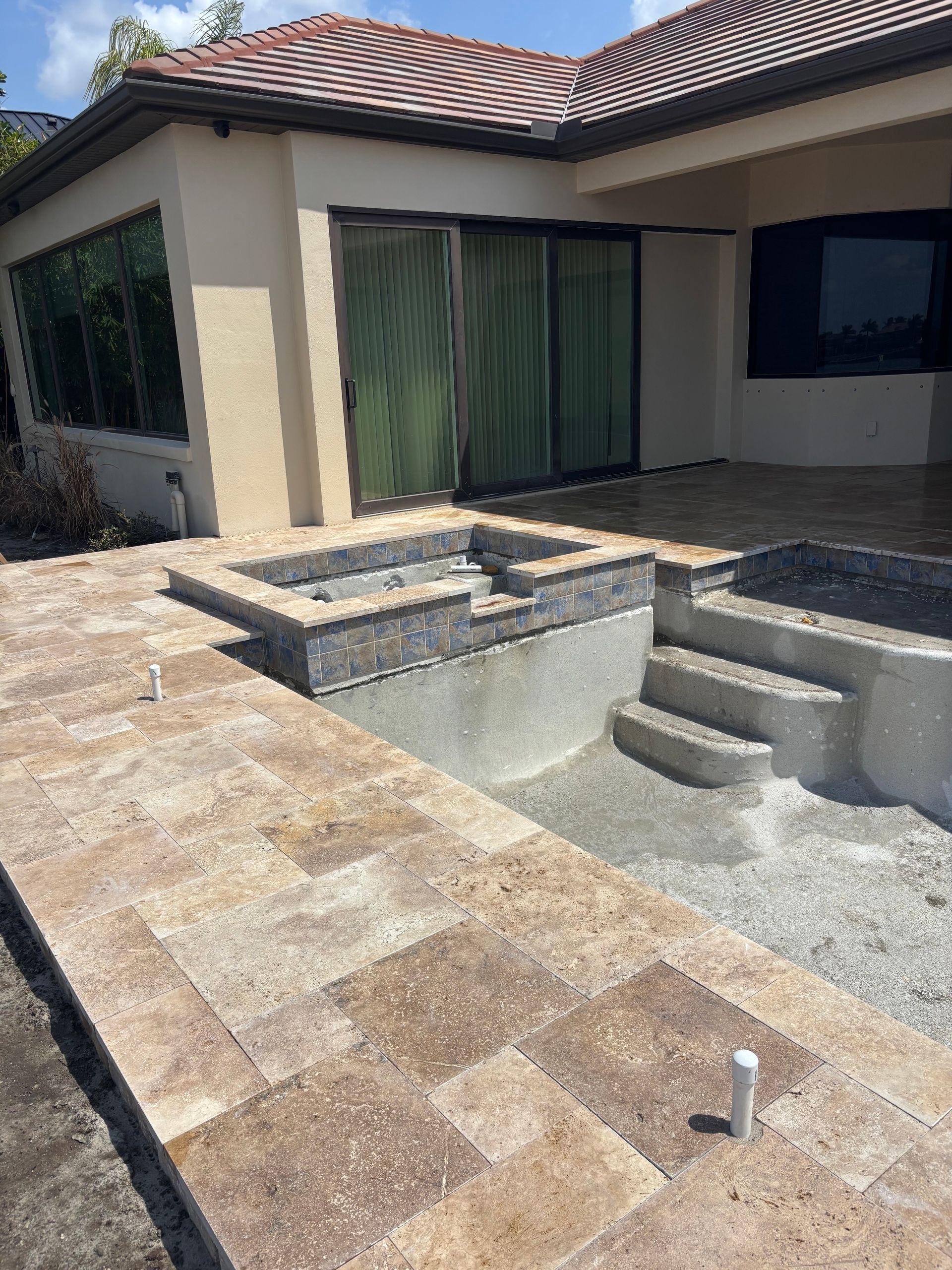 A patio with tan pavers, a partially finished concrete spa, and steps leading to a house with beige walls and a tile roof.
