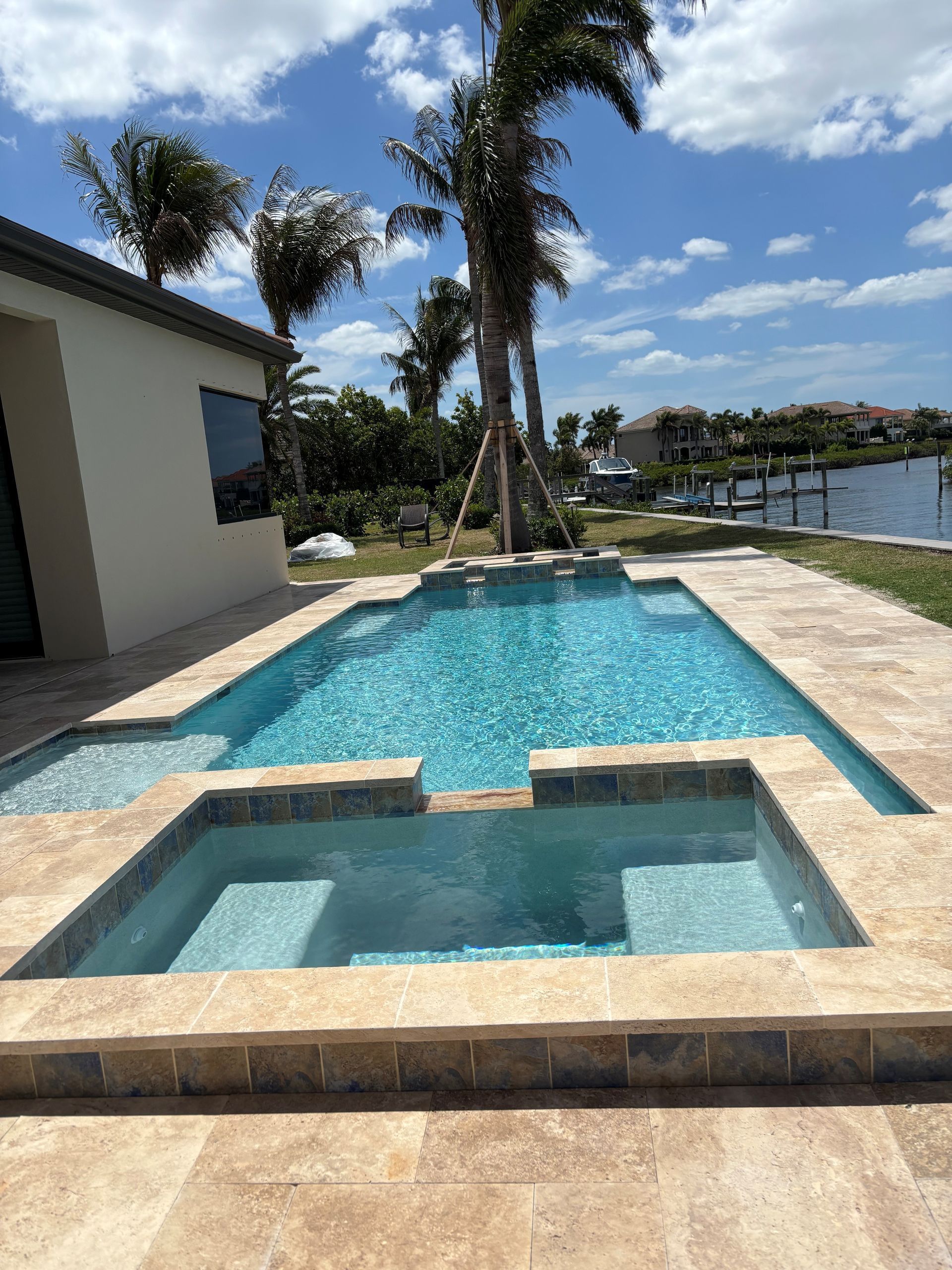 Patio pool with a small hot tub, turquoise water, palm trees, and a waterfront view under a blue sky.