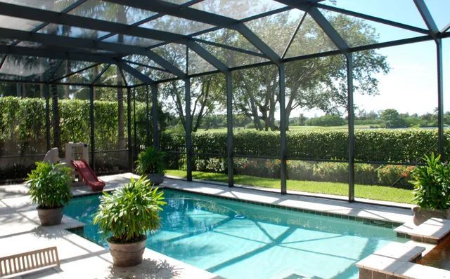 Covered backyard pool with lounge chairs, potted plants, and a sunlit view of green hedges and trees