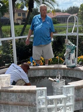 Two men working on a circular stone fountain in a garden area, one standing and one leaning over the basin
