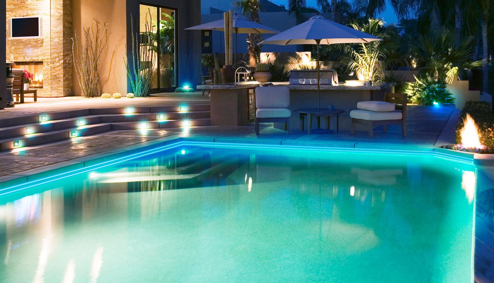 Illuminated backyard pool at night with turquoise water, patio seating, and warm house lights