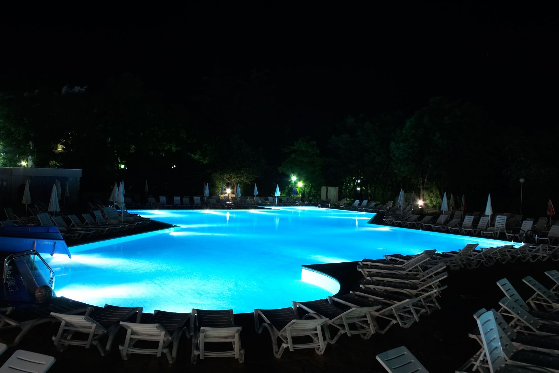 Illuminated blue swimming pool at night, surrounded by lounge chairs and dark trees