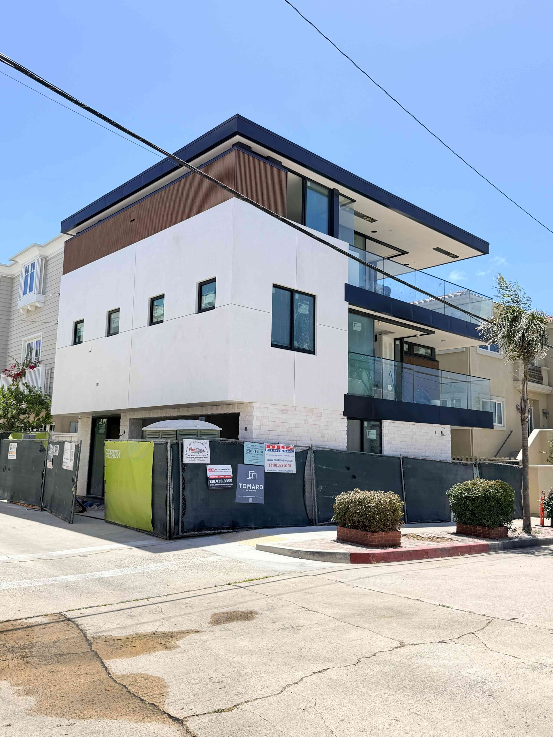 Modern three-story apartment building with balconies, white walls, and a street-facing trash enclosure under a blue sky