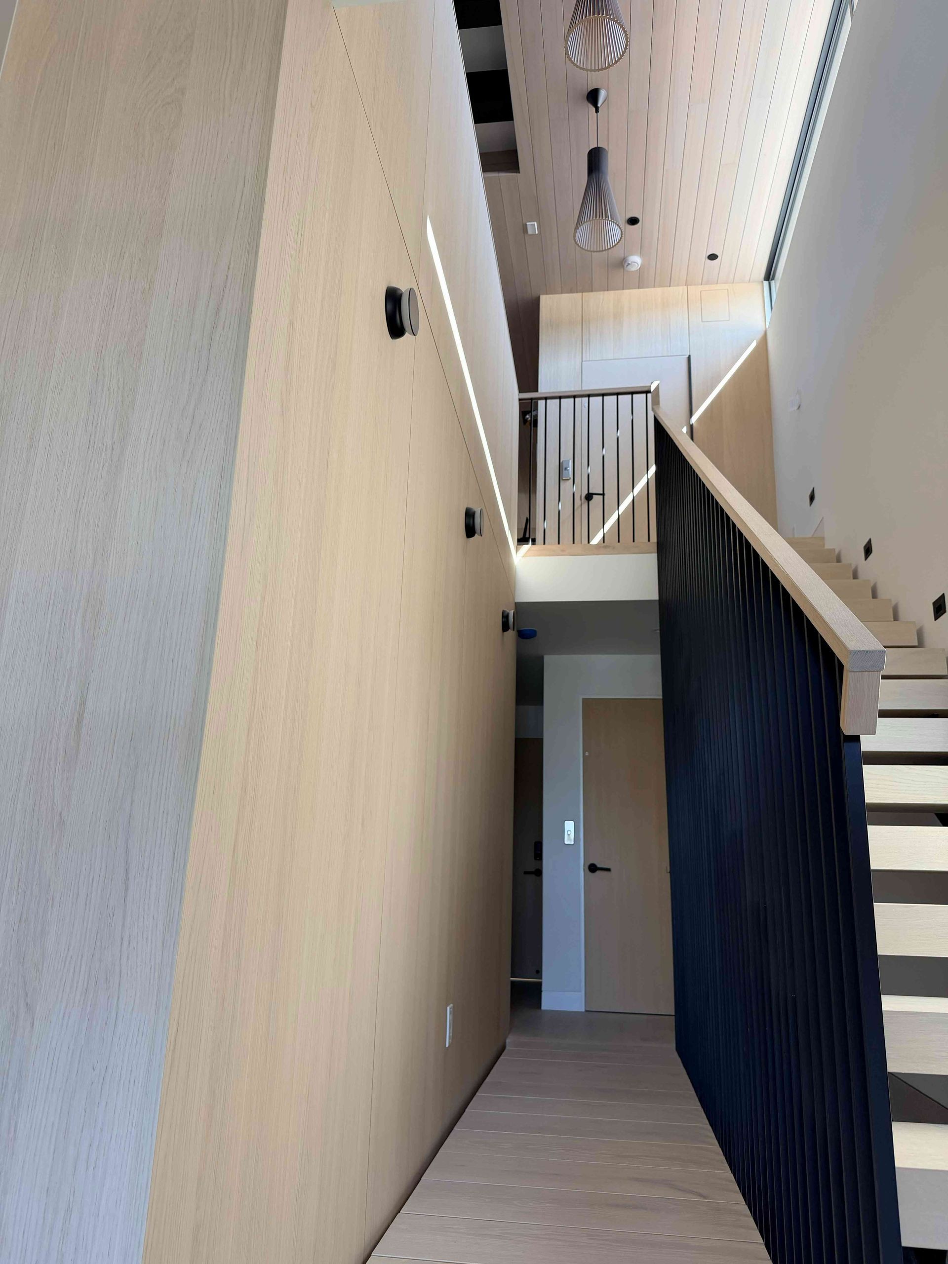 Modern interior hallway with high ceilings, beige walls, stair railing, and dark wood accent paneling.