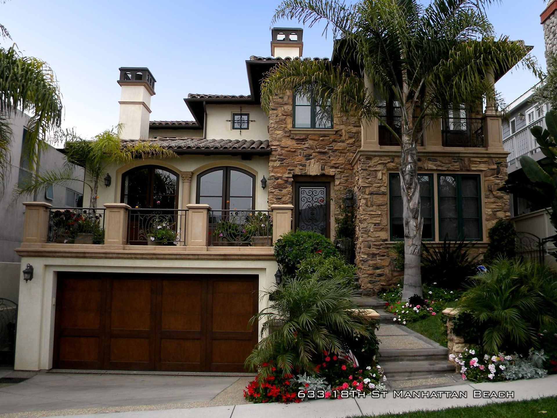 Large stone-and-stucco house with a wooden garage, arched entry, and lush front landscaping