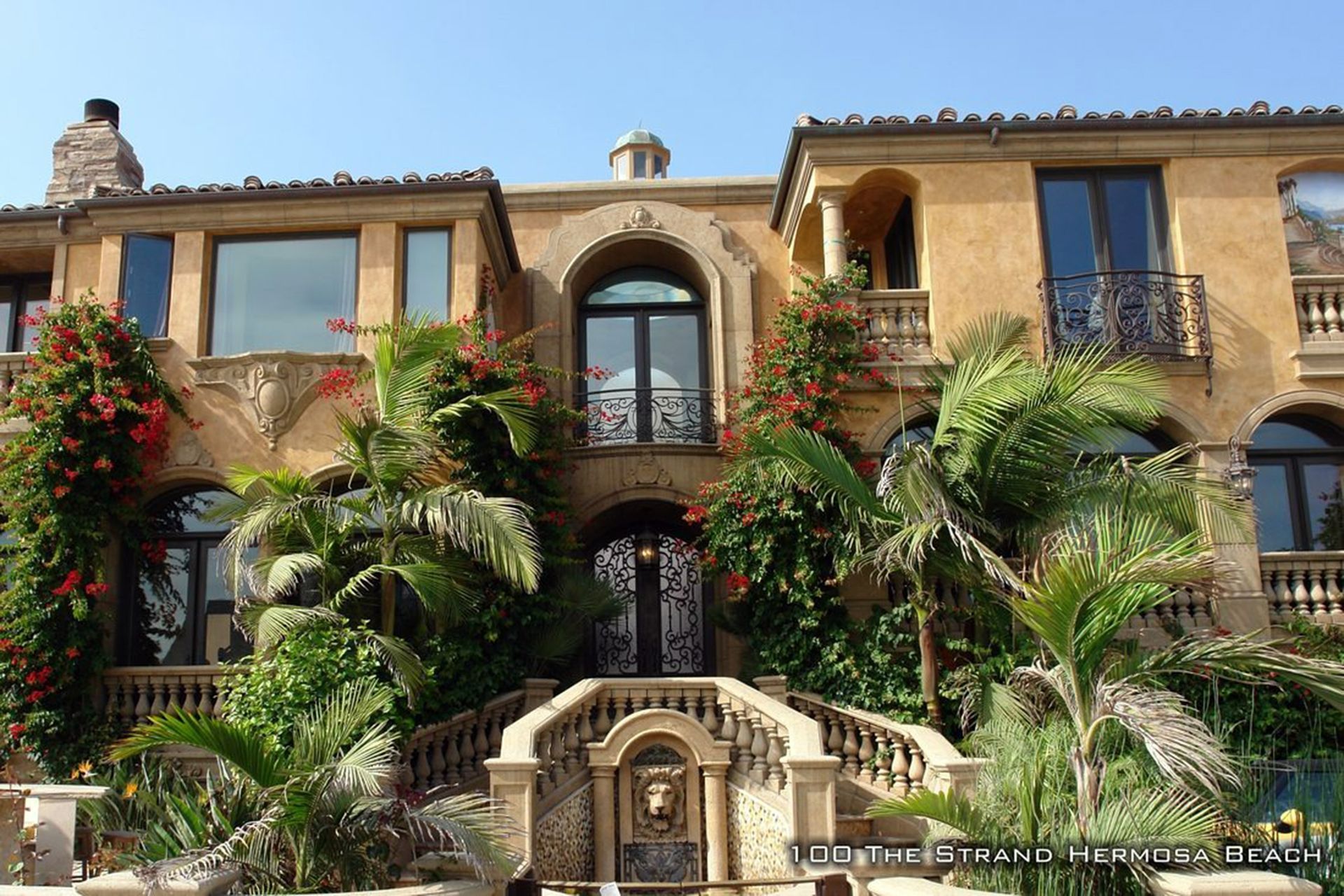 Mediterranean-style villa with arched balcony, palm trees, and lush flowering plants in front