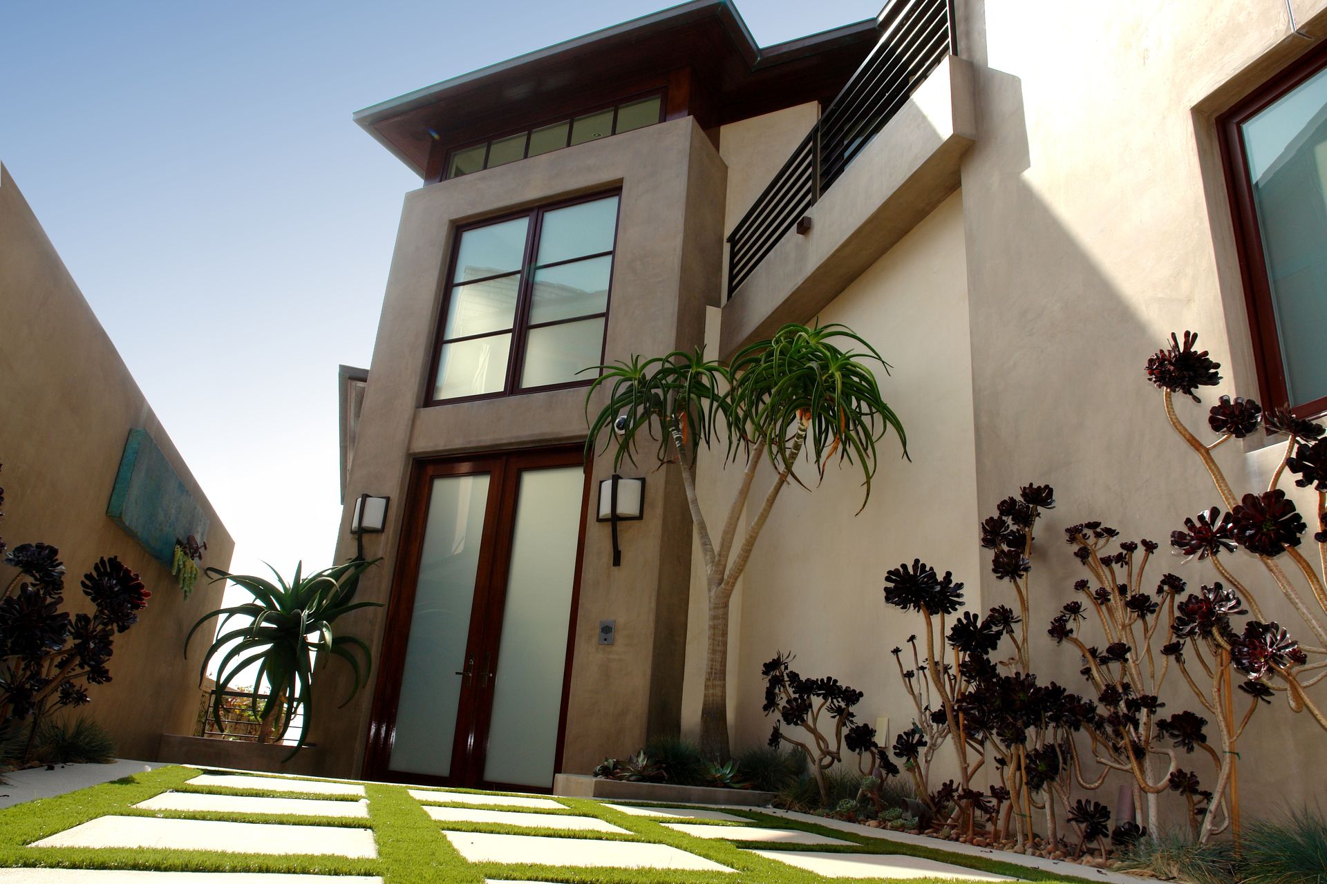 Modern two-story house with large windows, beige walls, and a landscaped front yard with stepping stones