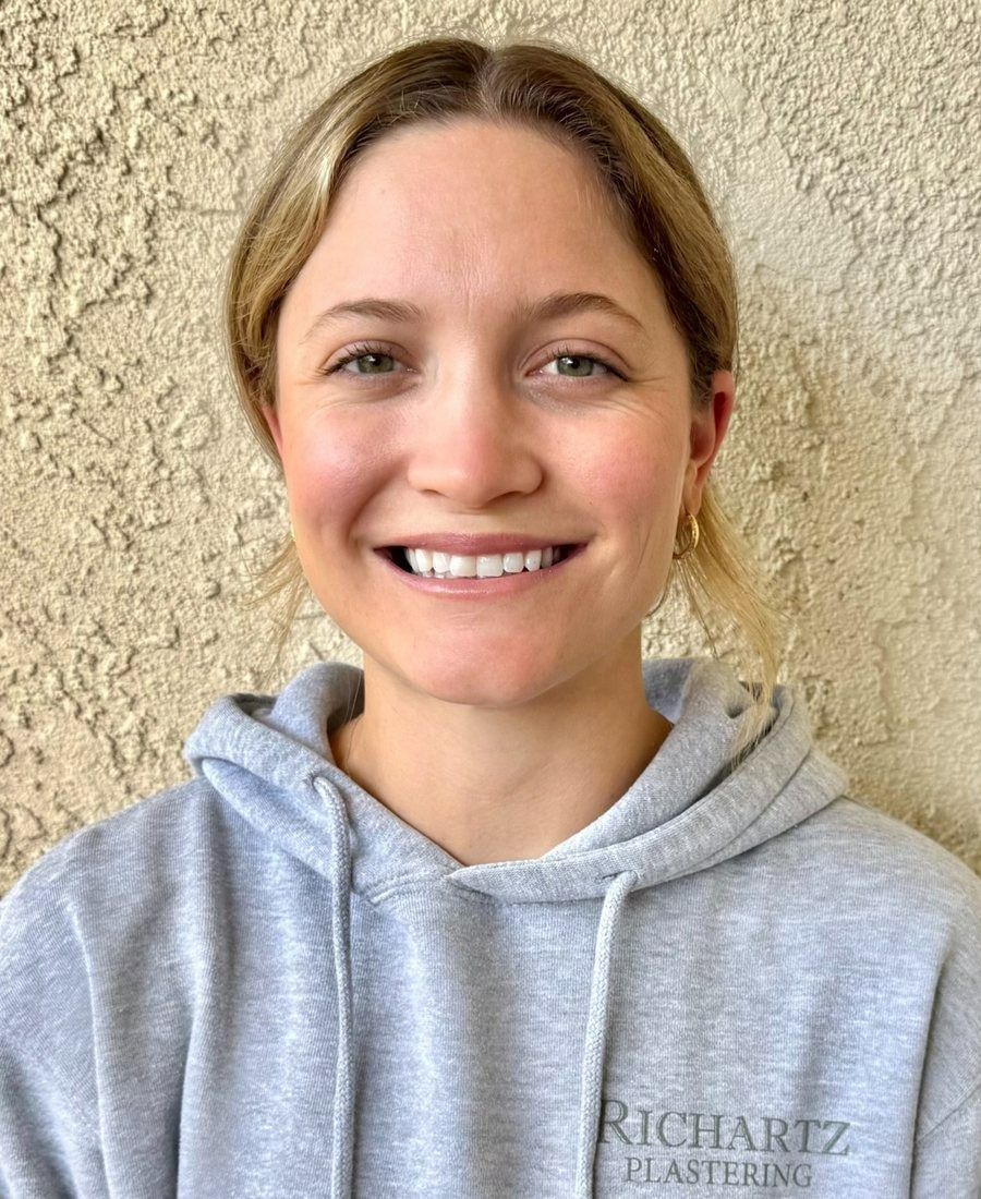 Smiling person in a gray hoodie standing against a textured beige wall.