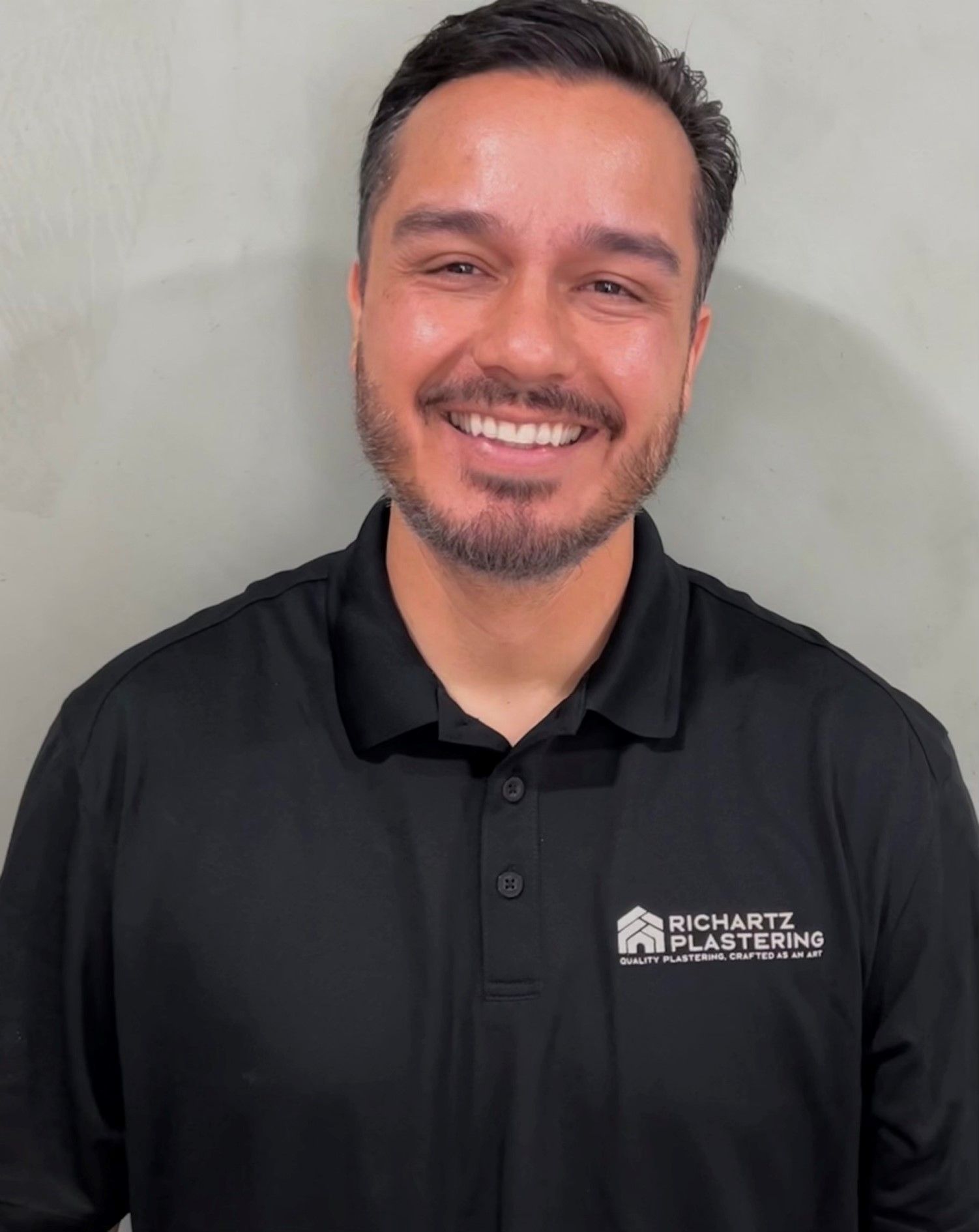 Smiling man in a black polo shirt with a company logo, standing against a light gray wall.