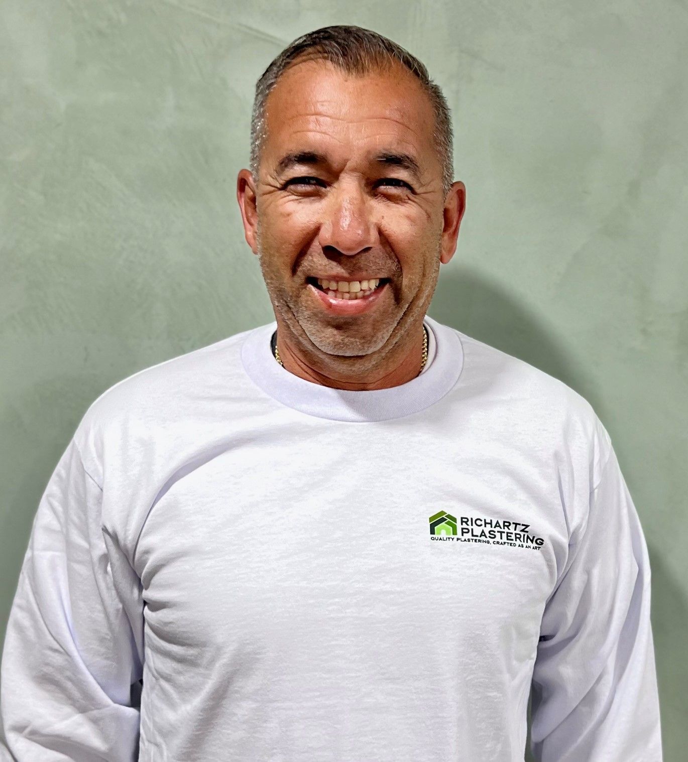 Smiling man in a white long-sleeve shirt standing against a plain green-gray background