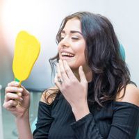 A woman is smiling while looking at her teeth in a mirror.