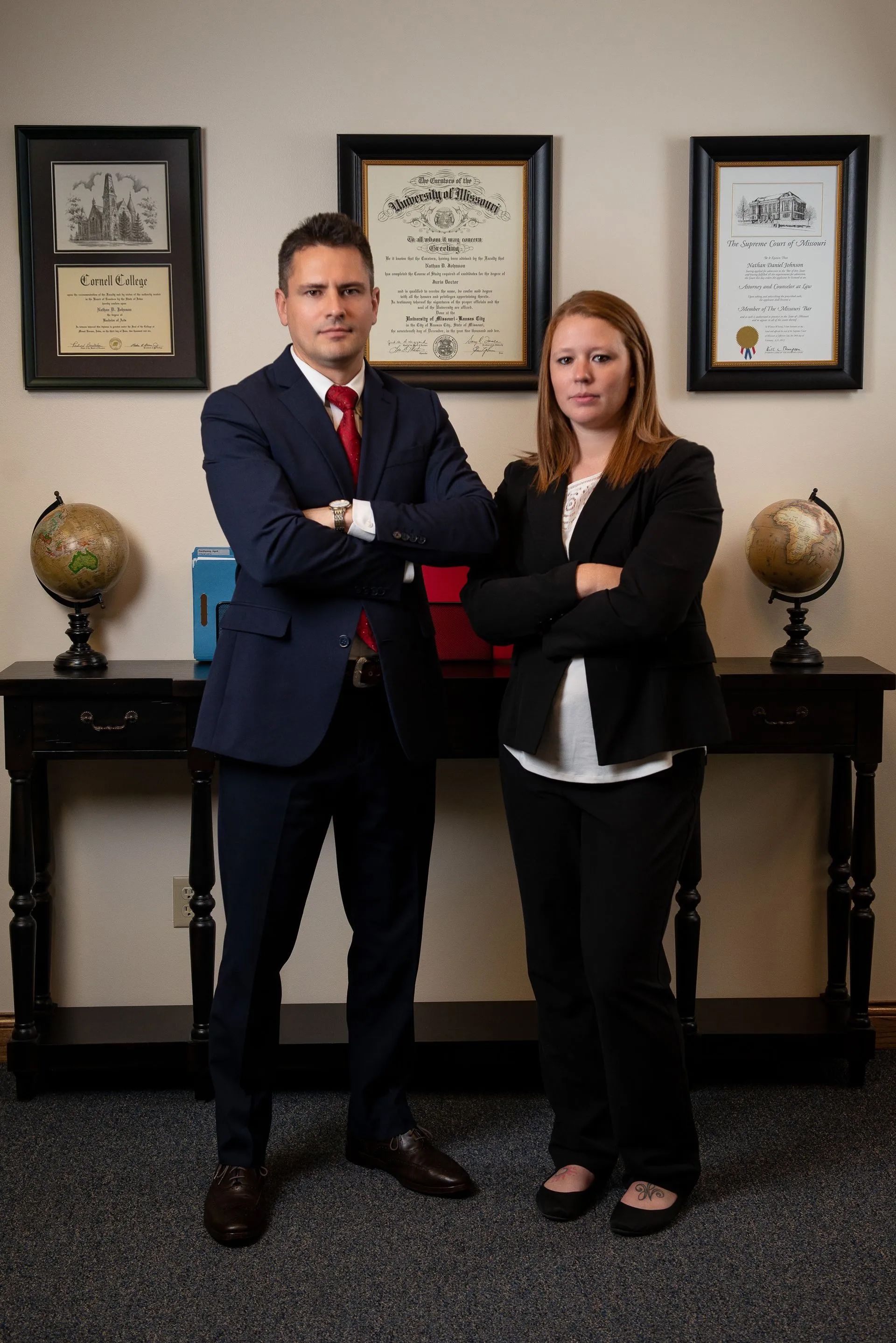Two professionals standing in an office, arms crossed, with certificates and globes on the wall behind them.