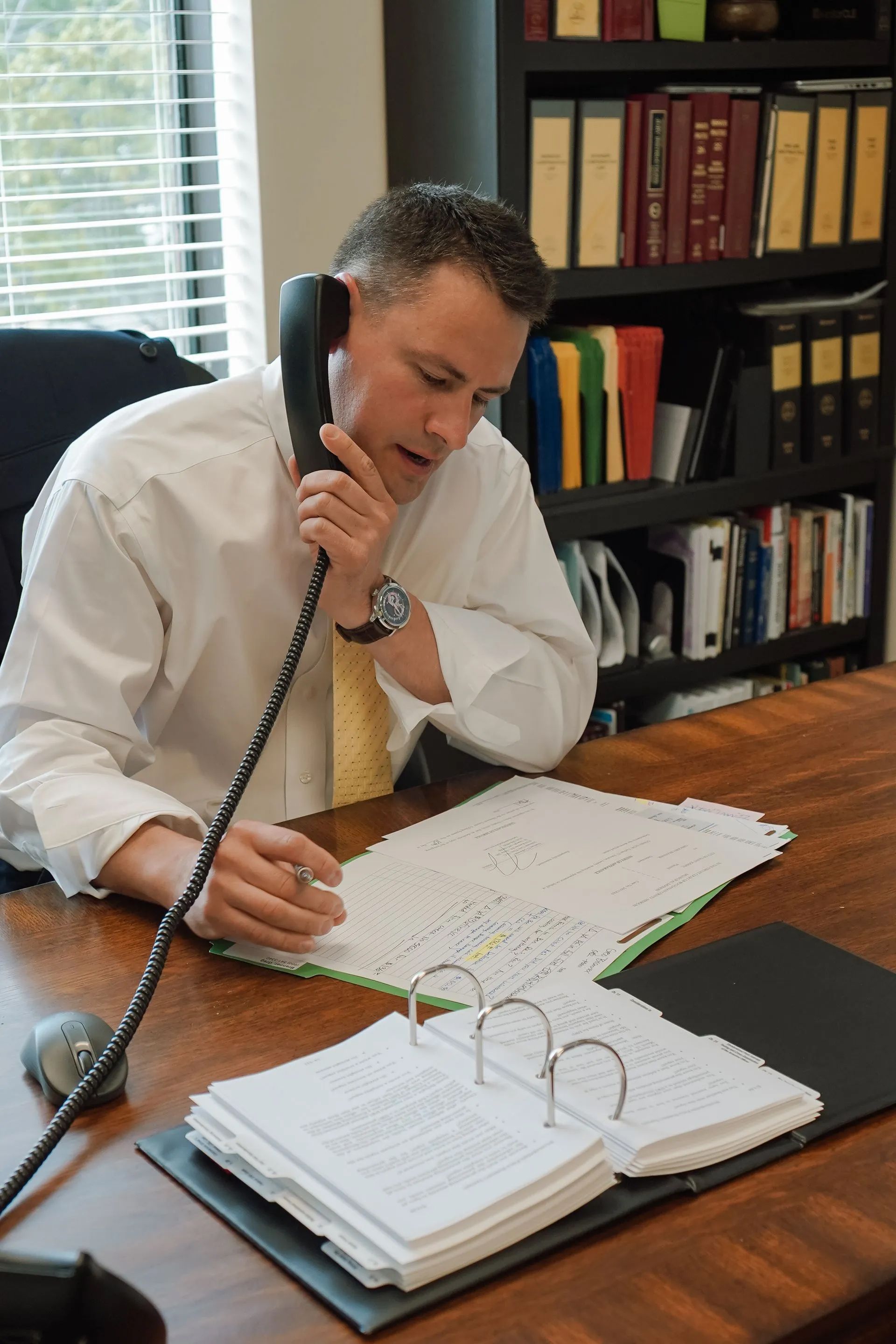 Man on a corded phone at a wooden desk, writing notes beside an open binder in an office.