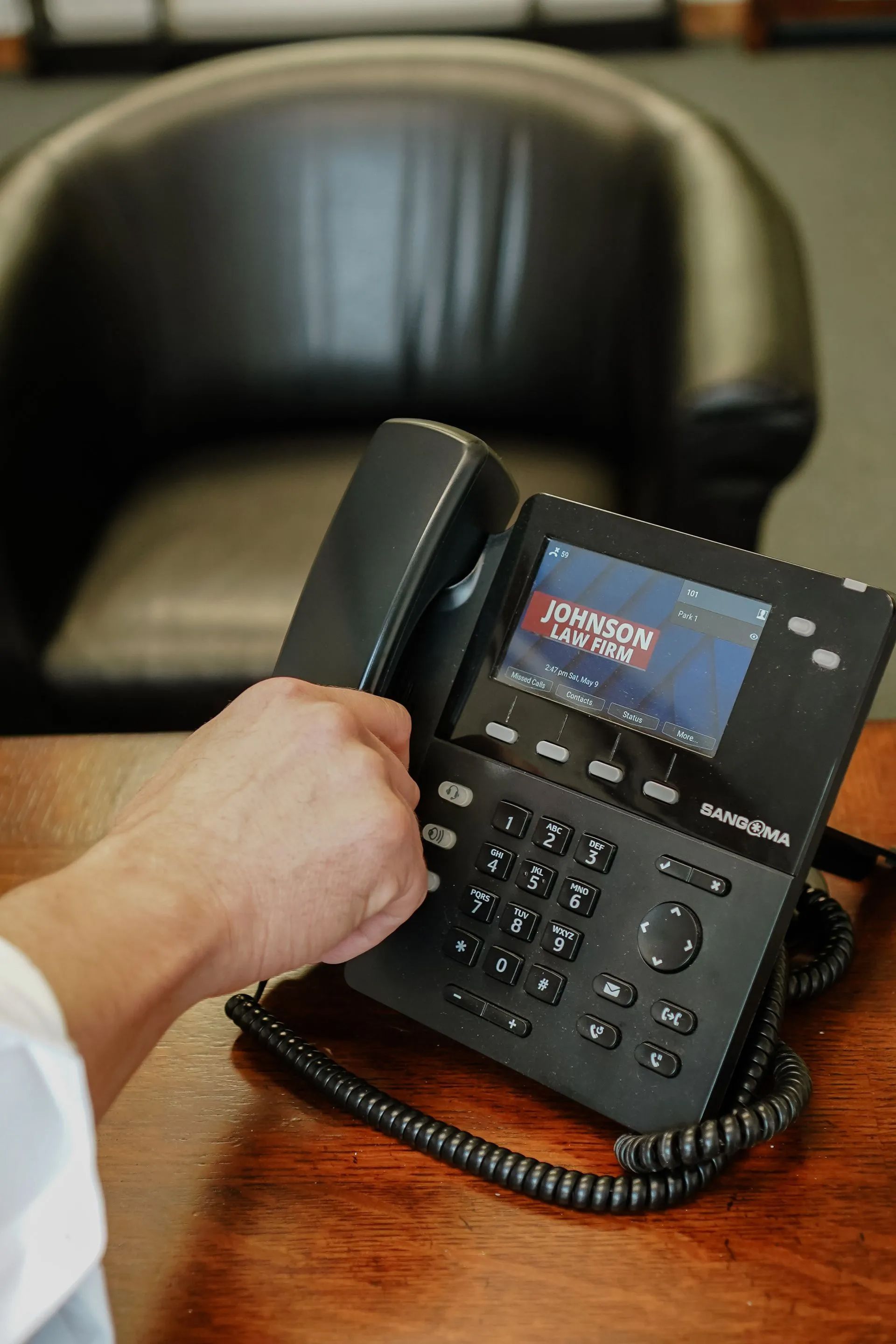 Hand holding a black office desk phone on a wooden table, with a leather chair in the background