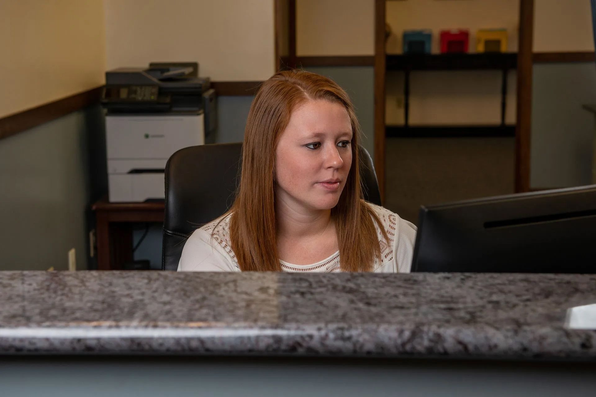 Woman seated at a desk in an office, looking to the side past a computer monitor.