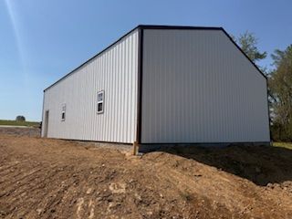 A white metal pole barn with dark trim stands on a dirt slope under a clear blue sky.  