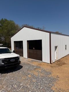A white metal garage building with two brown doors and a side entry door, parked next to a white pickup truck on gravel. 