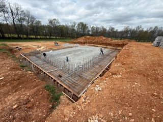 Workers install rebar and plumbing for a building foundation in a large dirt excavation site.
