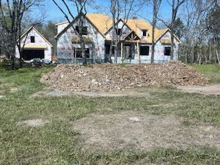 A house under construction with exposed wooden framing and white siding, sitting behind a large pile of dirt in a field. 